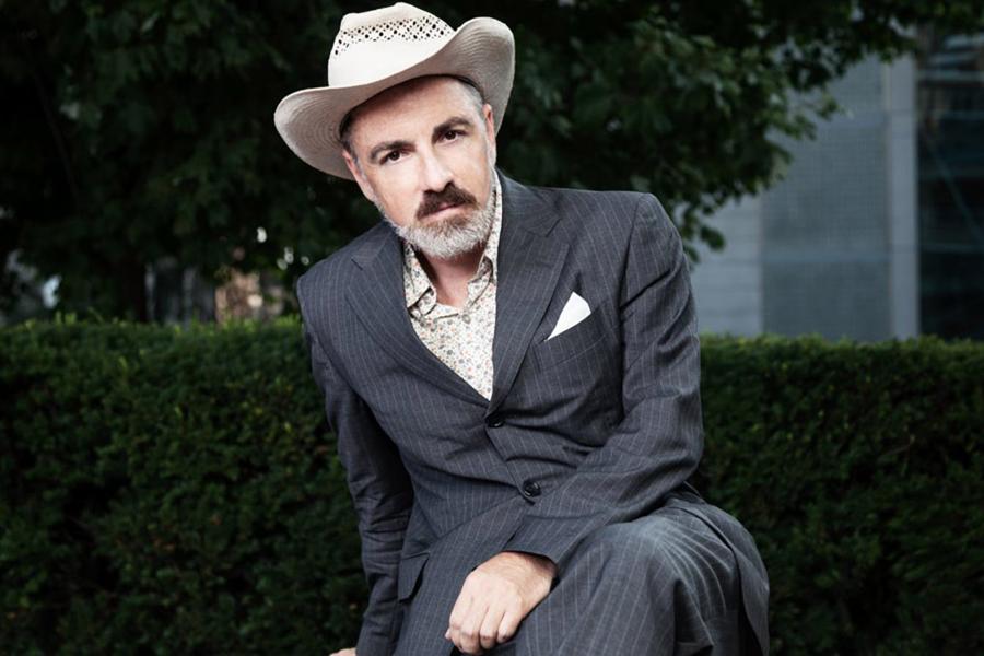 a publicity portrait of Howard Fishman, sitting on a park bench, in a grey suit and a stetson cowboy hat