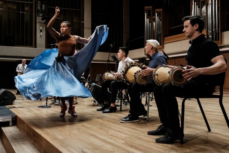 A woman in a blue skirt dancing on a stage in a well lit space in front of 3 seated men playing hand drums.. 