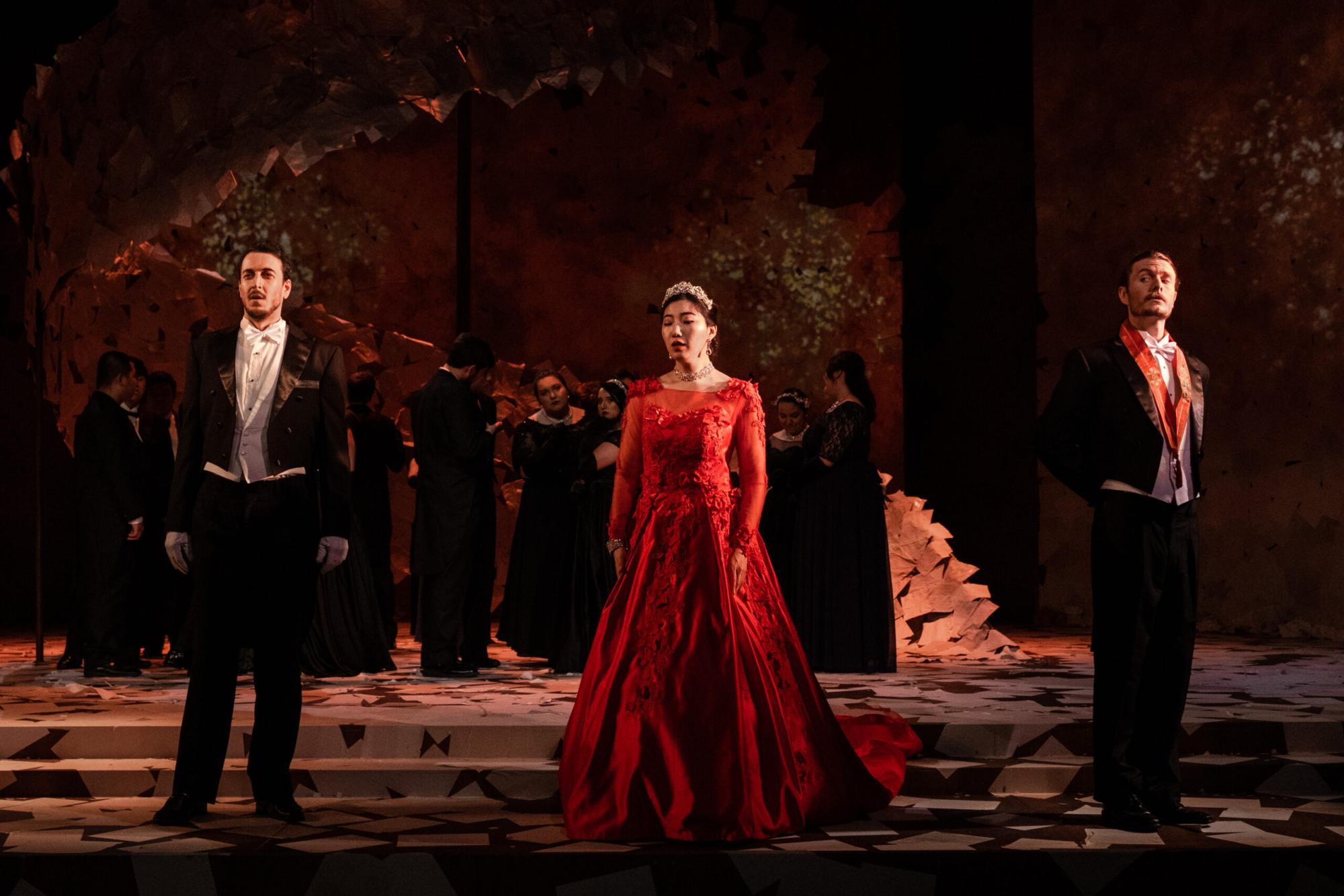 A sad woman in a red dress on stage, flanked by two men in tuxedos.