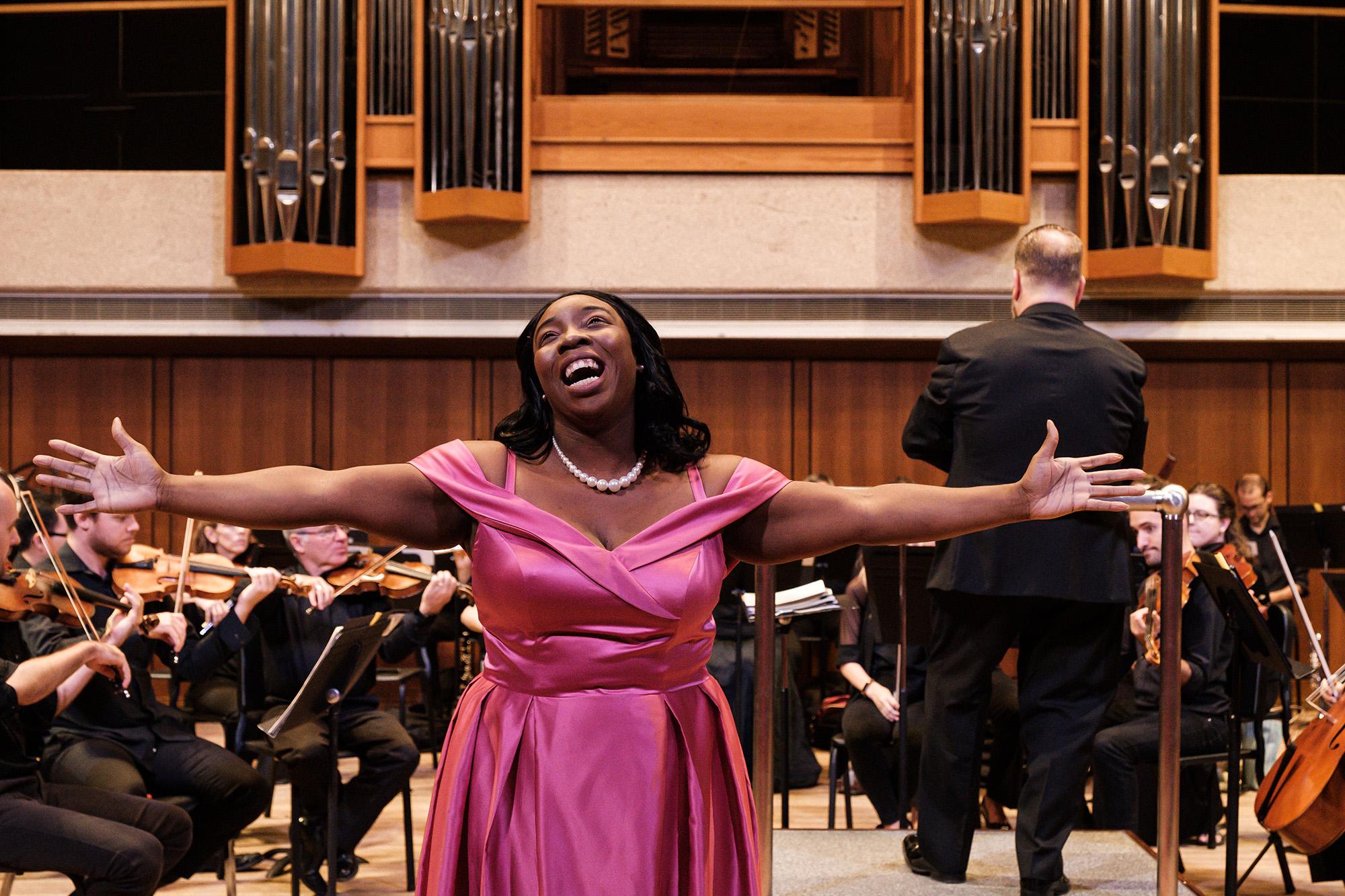 A black woman wearing a pink gown spreads her arms while singing. She stands in front of an orchestra.