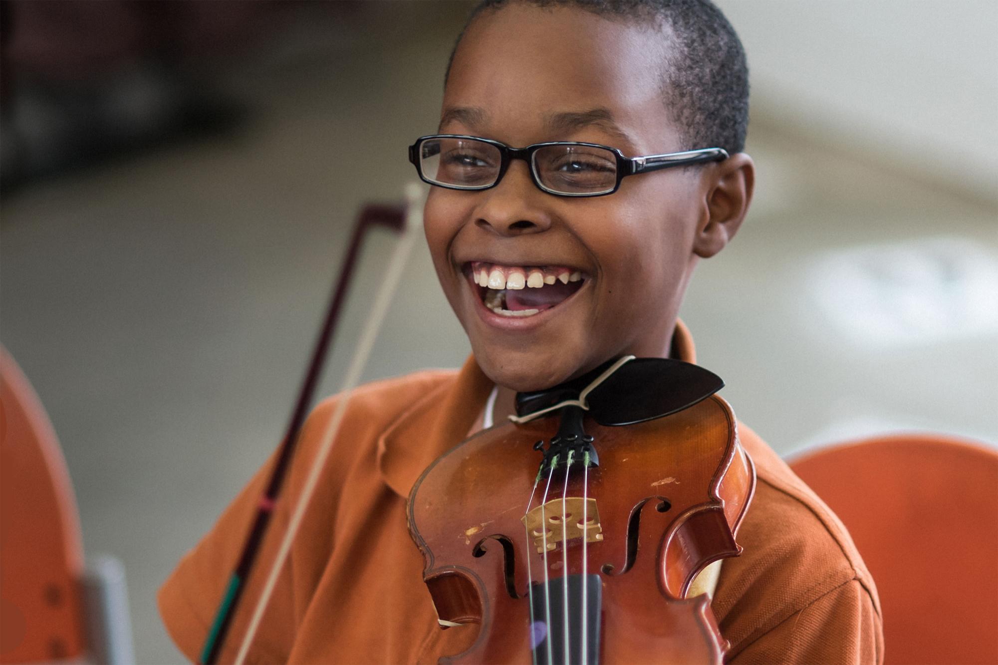 A young violin student holding his instrument in playing position, laughs gleefully during a music lesson