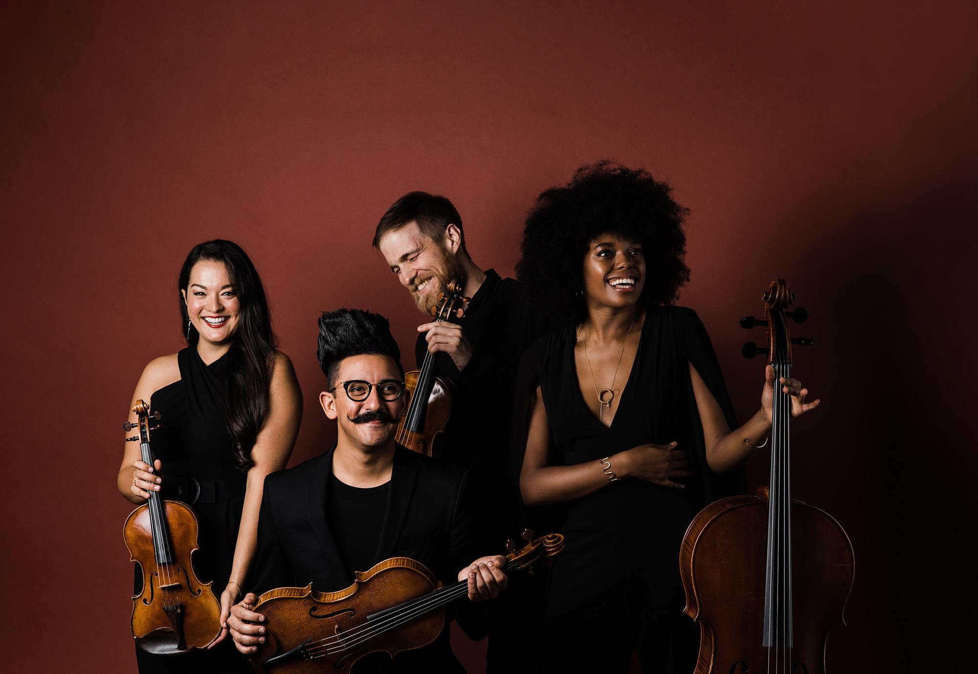 Thalea String Quartet pose with instruments in front of a red wall