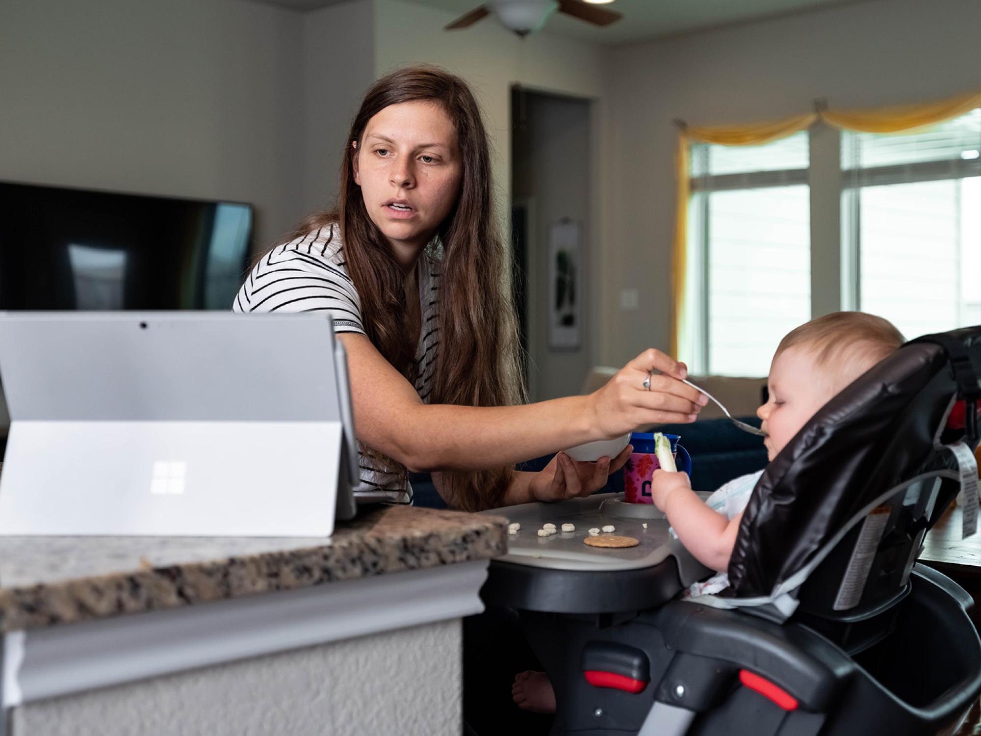 Heather sits at a barstool in her kitchen, feeding Daisy who is in a high char.  Her laptop is on a nearby countertop, and Heather is looking at the screen.  