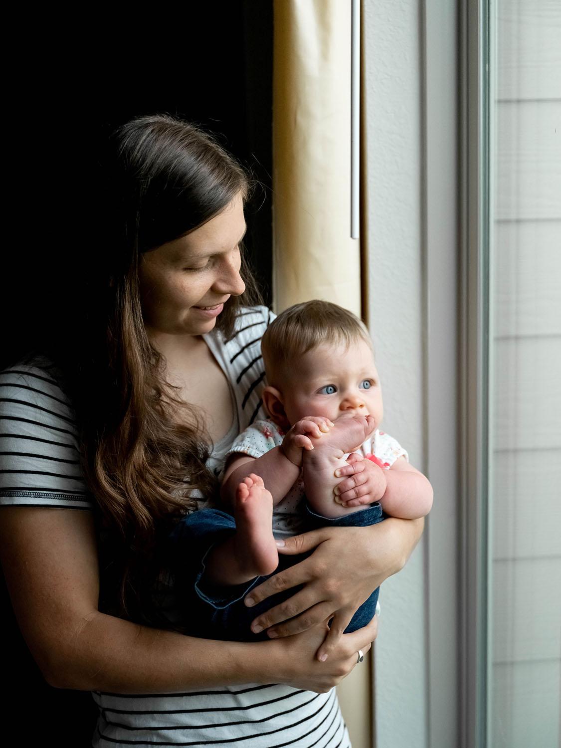 A woman stands near a window looking down at a toddler in her arms .The toddler looks out the window, holding her foot to her face.