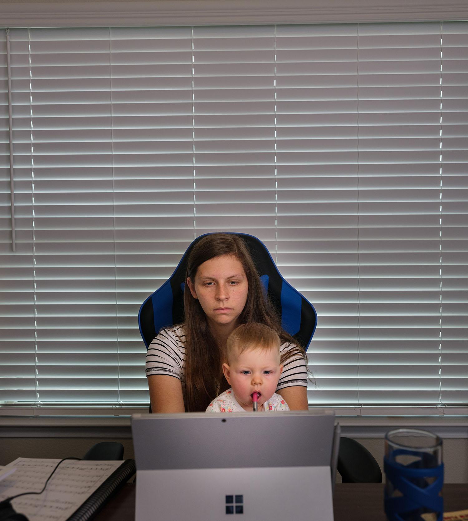 Heather sits at her desk, attending a class on her laptop. Her daughter sits on her lap, with a lollipop in her mouth.