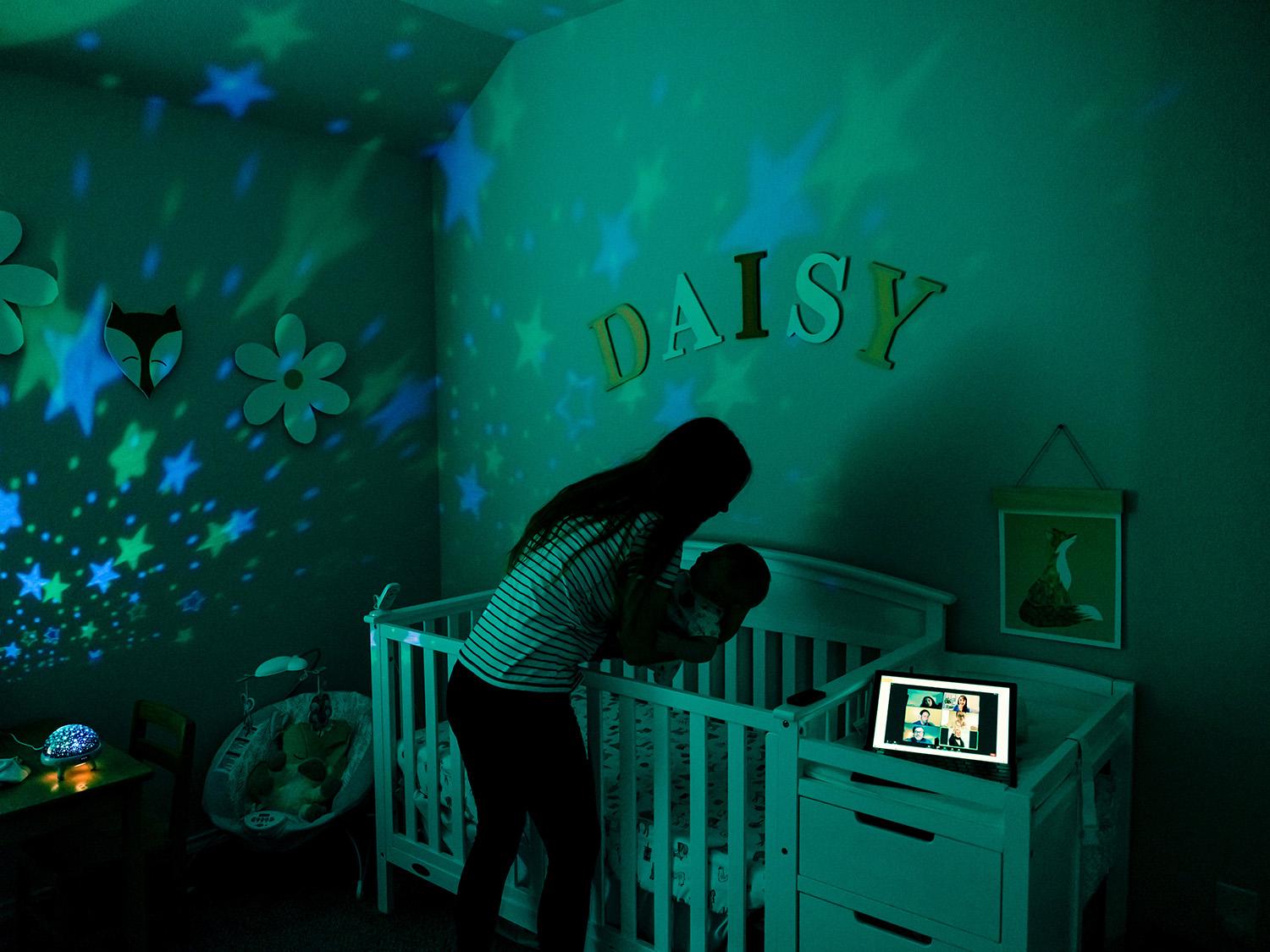 Heather stands over a crib in a dark nursery, putting Daisy down to sleep.  To the side of the crib, her laptop is logged in to a virtual classroom where a discussion is taking place.