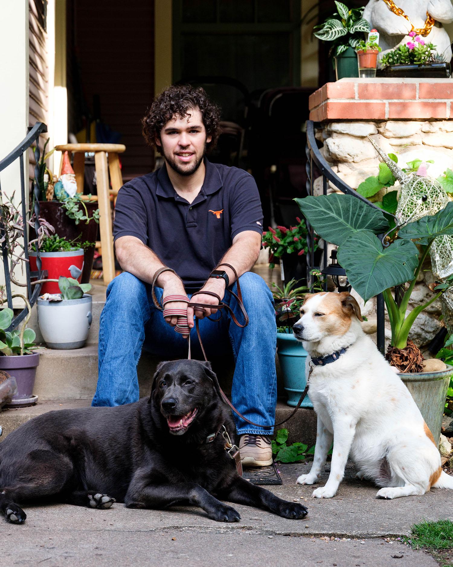 Nick, A student in a UT shirt sits on the front steps of his home, with his two dogs at his side.