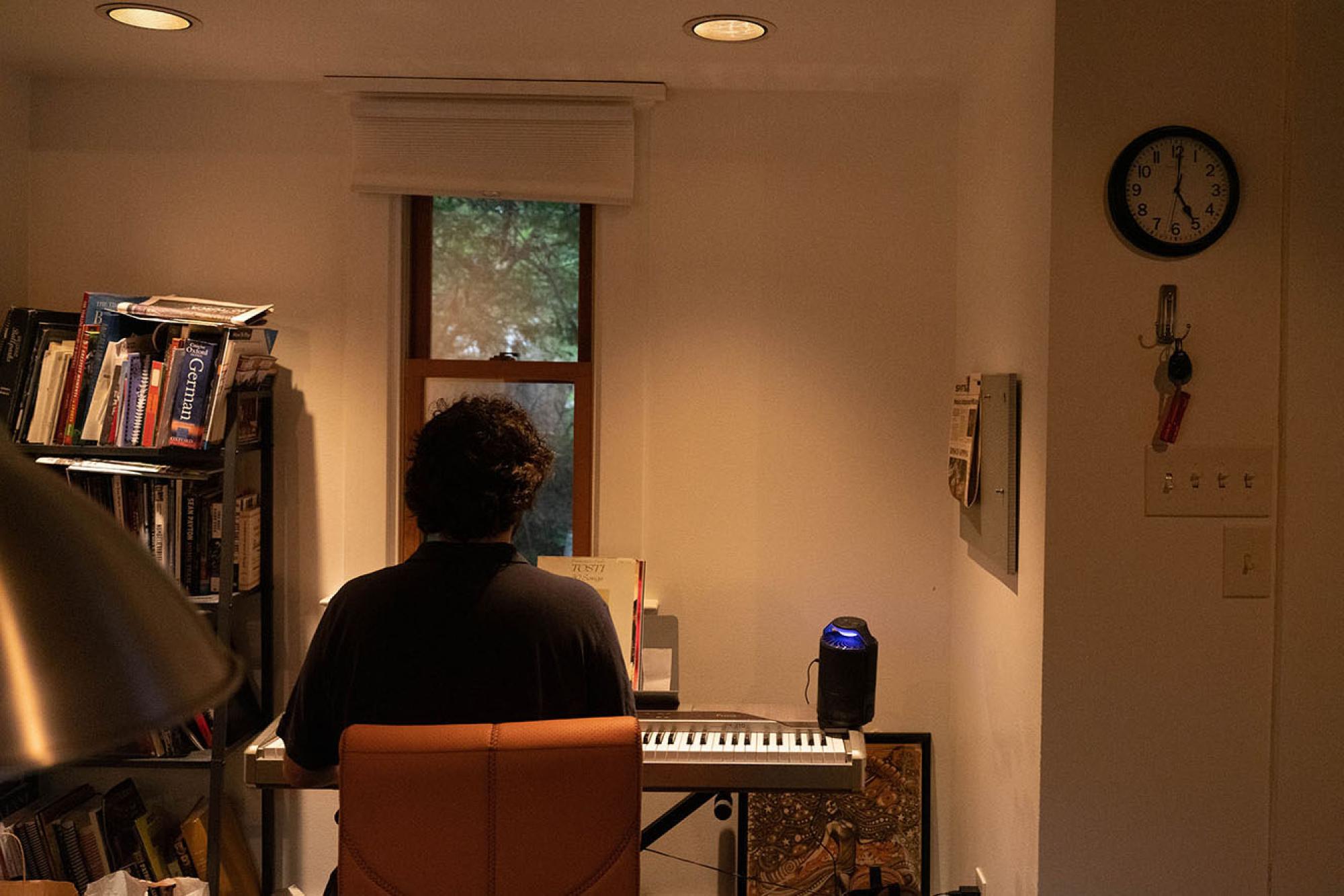 A Man sits alone in his apartment, at an electronic keyboard with his back to the camera practicing music. 