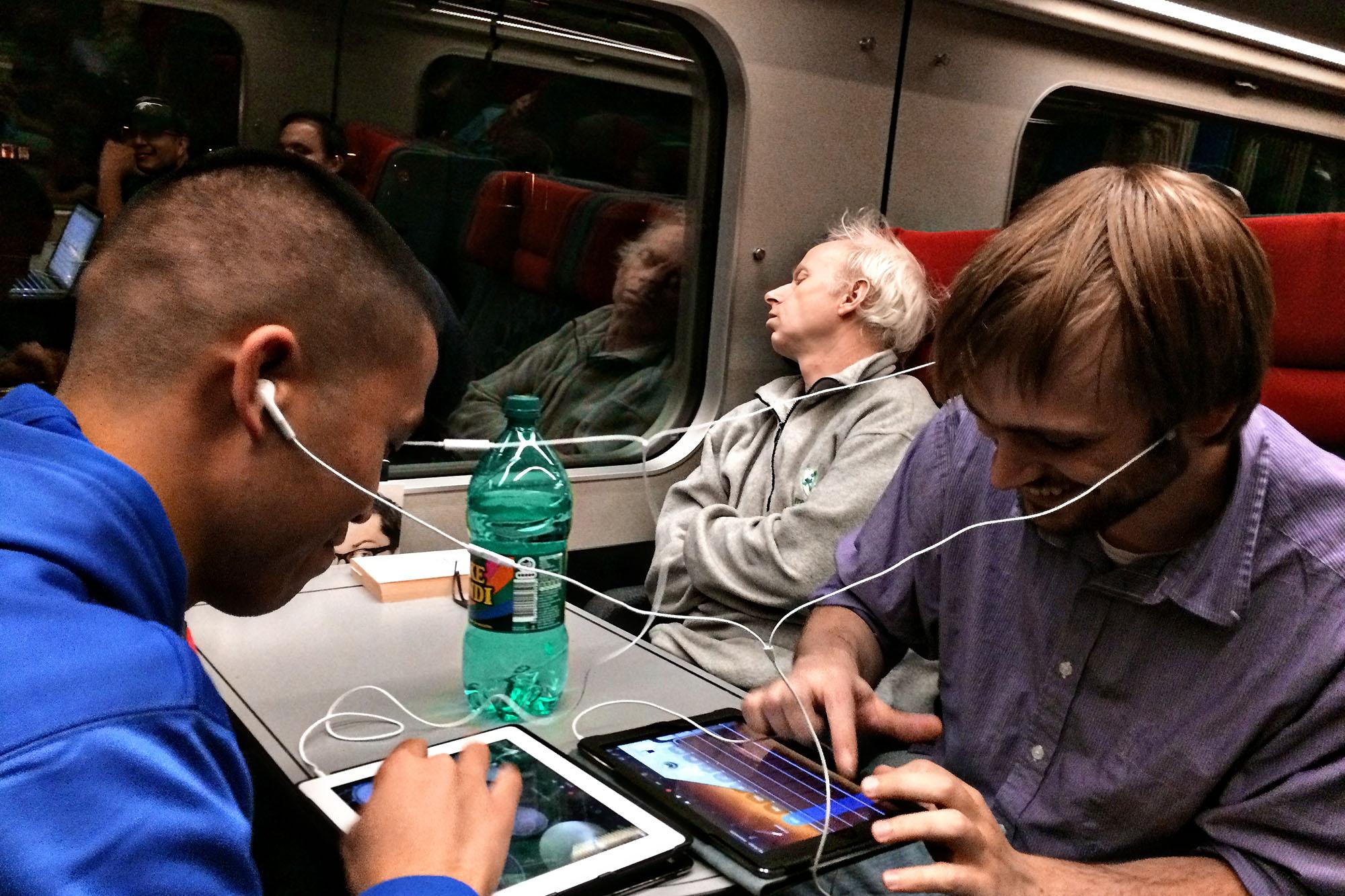 Students playing a game on tablets while on a train.  A man is sleeping in his train seat the background 