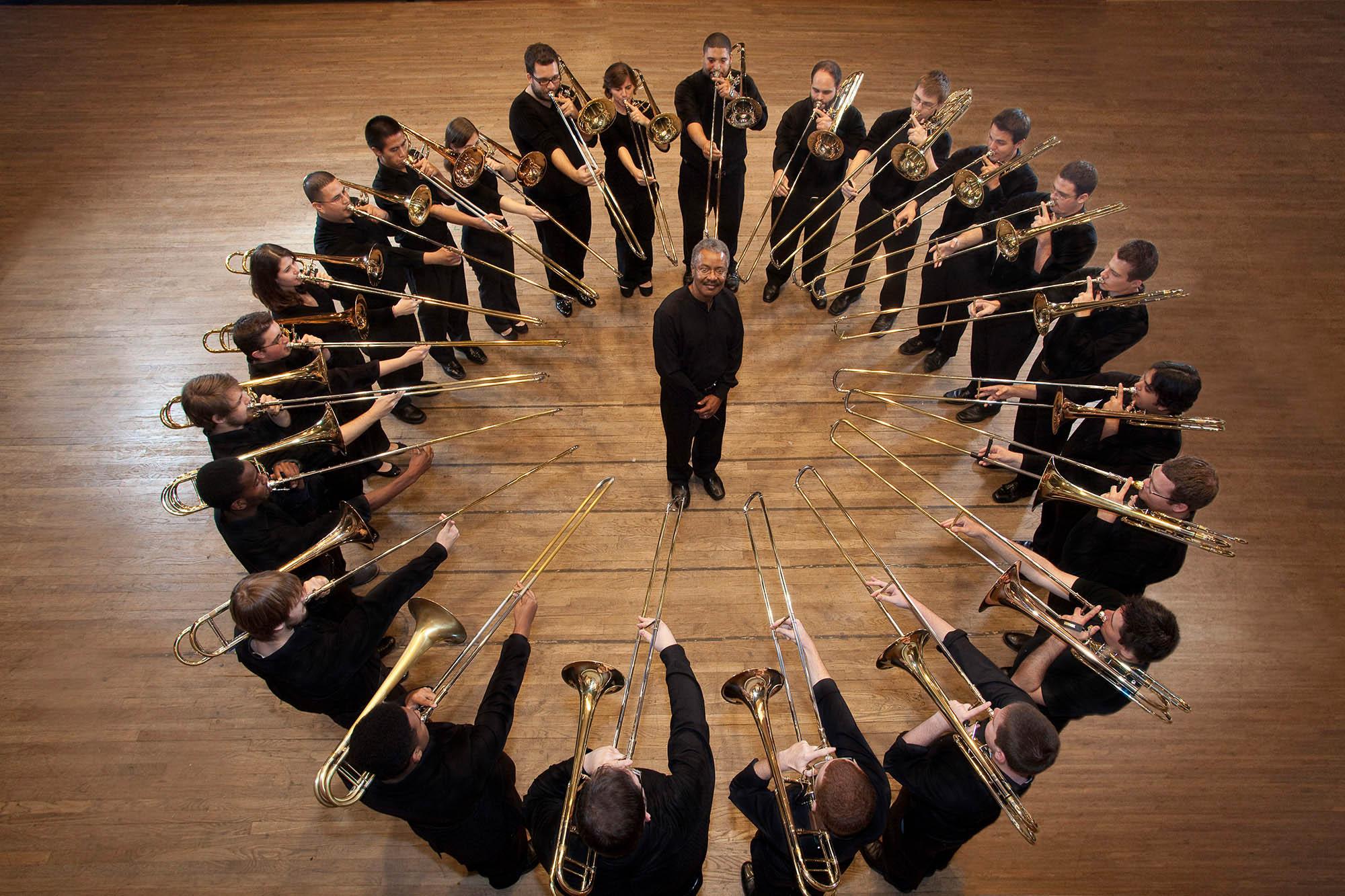 Trombone choir stand in a circle on the Bates Recital stage, with their horns raised as if to play. Nathaniel Brickens stands in the center of the circle looking the camera.