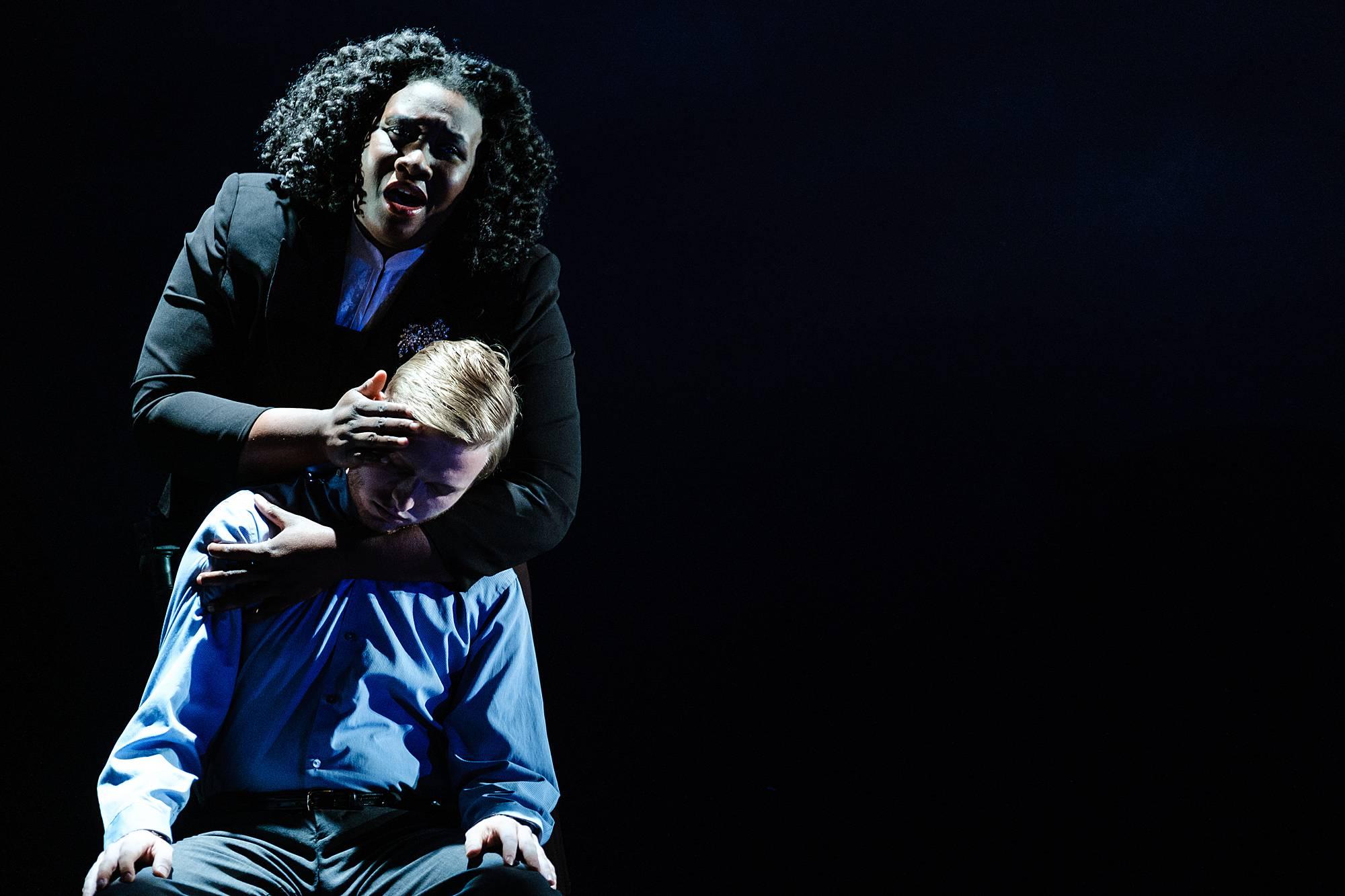 on a very dark stage, a man sits in a chair, looking to be in deep pain, as a woman stands behind him, cradling is face, singing in agony.