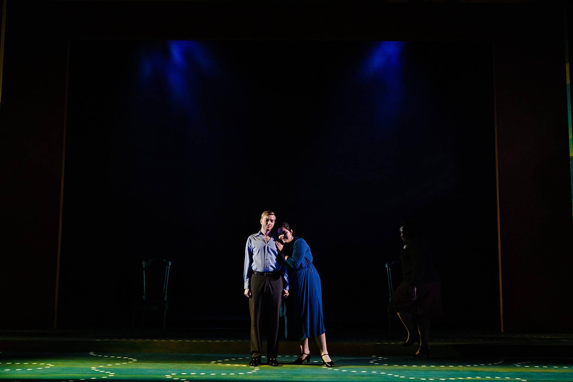 A man and woman stand together on a dark, expansive set, the man stares straight ahead with arms at his side, while the woman grips the man tightly, and rests her head on his shoulder