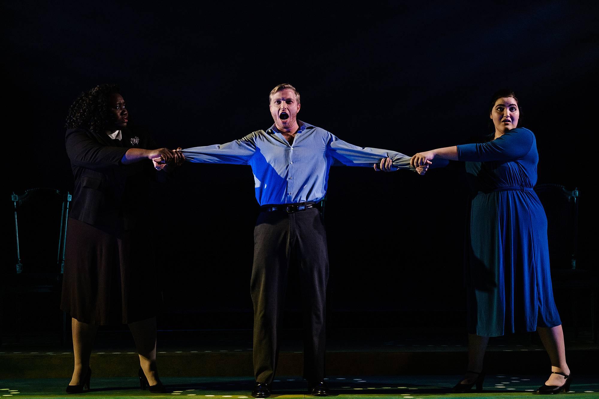 A man stands center stage, while two women on either side of him tug at his arms, as if they are fighting over him. 