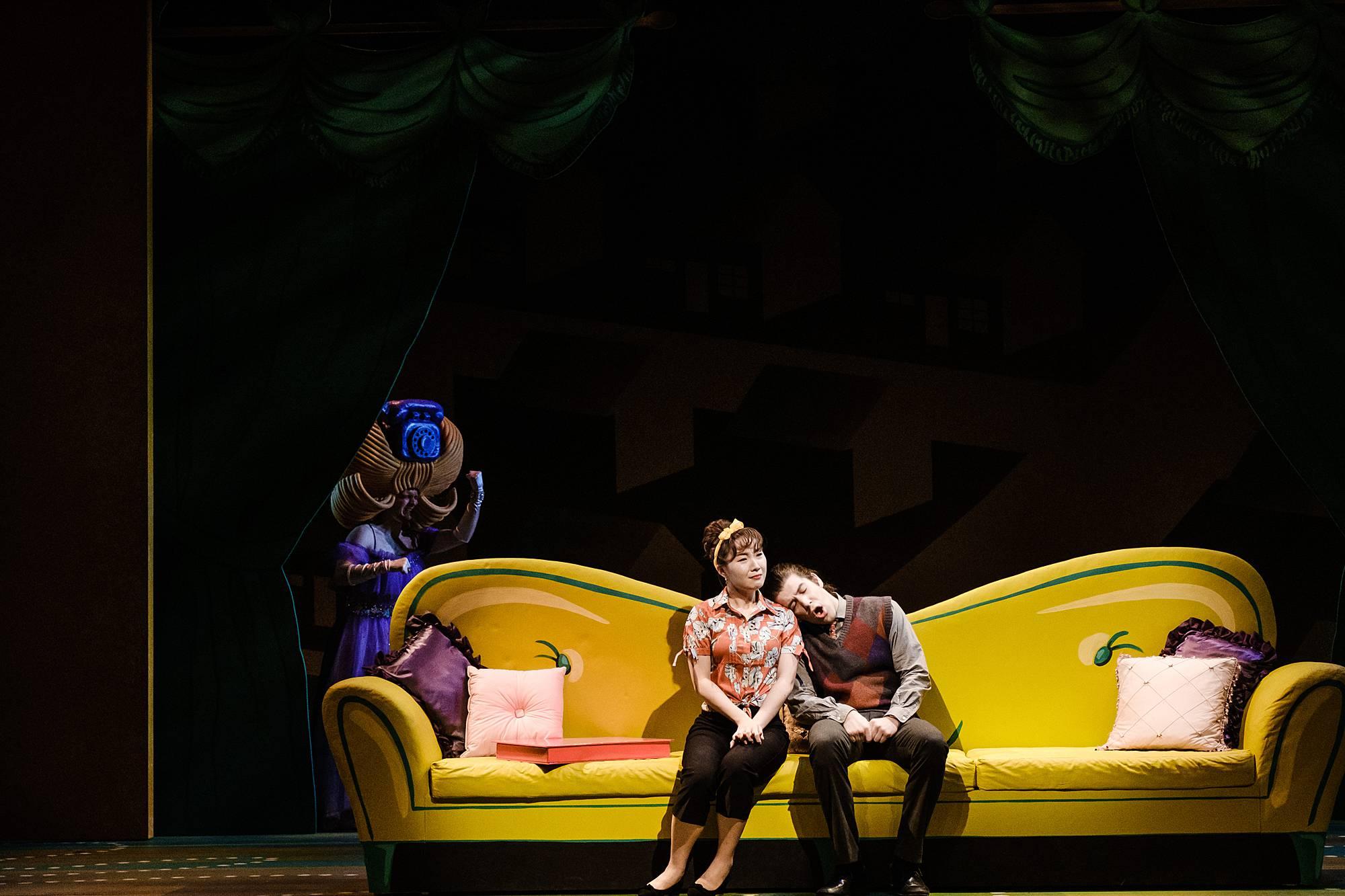 A woman and man sit on an overside cartoonish yellow couch, the man rests his head on the woman's shoulder.  A woman dressed in a foam bee-hive with with a telephone perched on top stands in the background. 