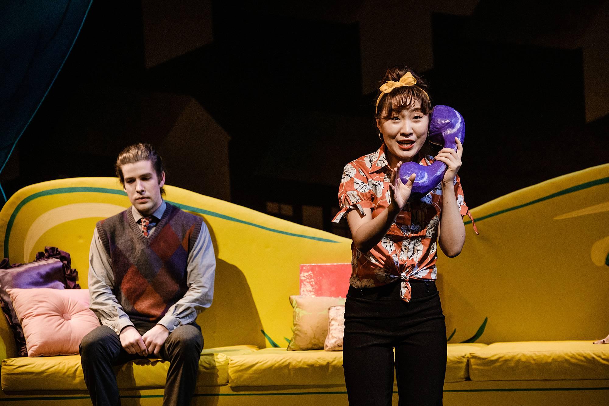 A woman stands center stage holding an oversized telephone receiver to her ear, listening, while a man sits on a large yellow couch behind her.