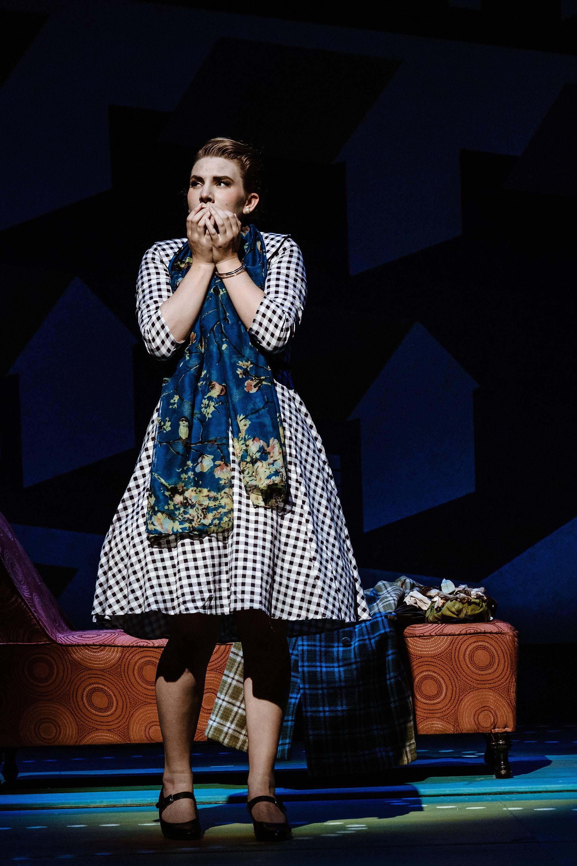 A woman stands in an overcoat and scarf, surrounded by suitcases, clutching her hands to her mouth nervously. 