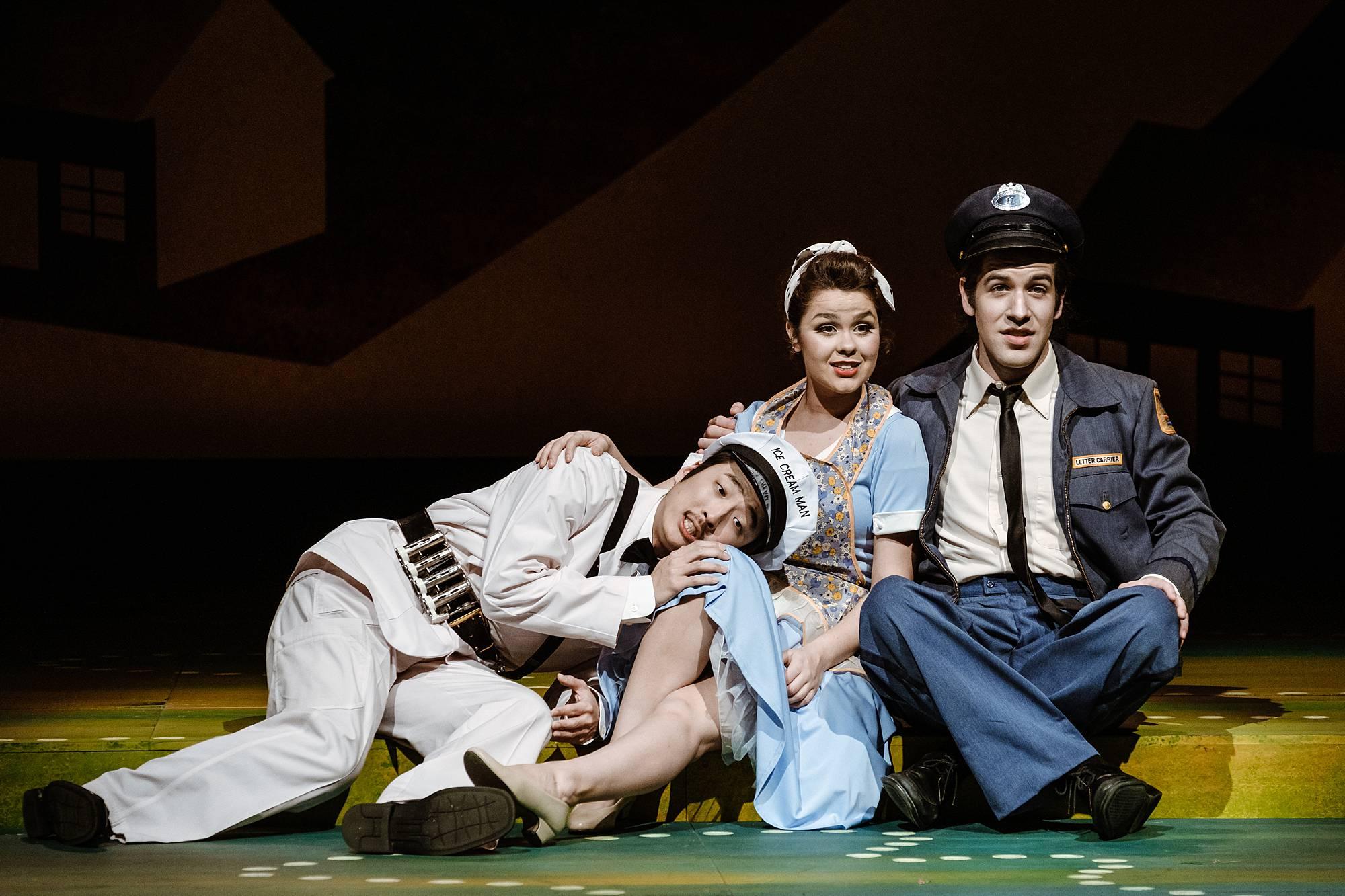 a man in a mid-century post office worker's uniform sits on the stage floor beside a woman in a maid's uniform. A man in a milkman's uniform lays on stage next to them with his head cradled in the woman's lap. 