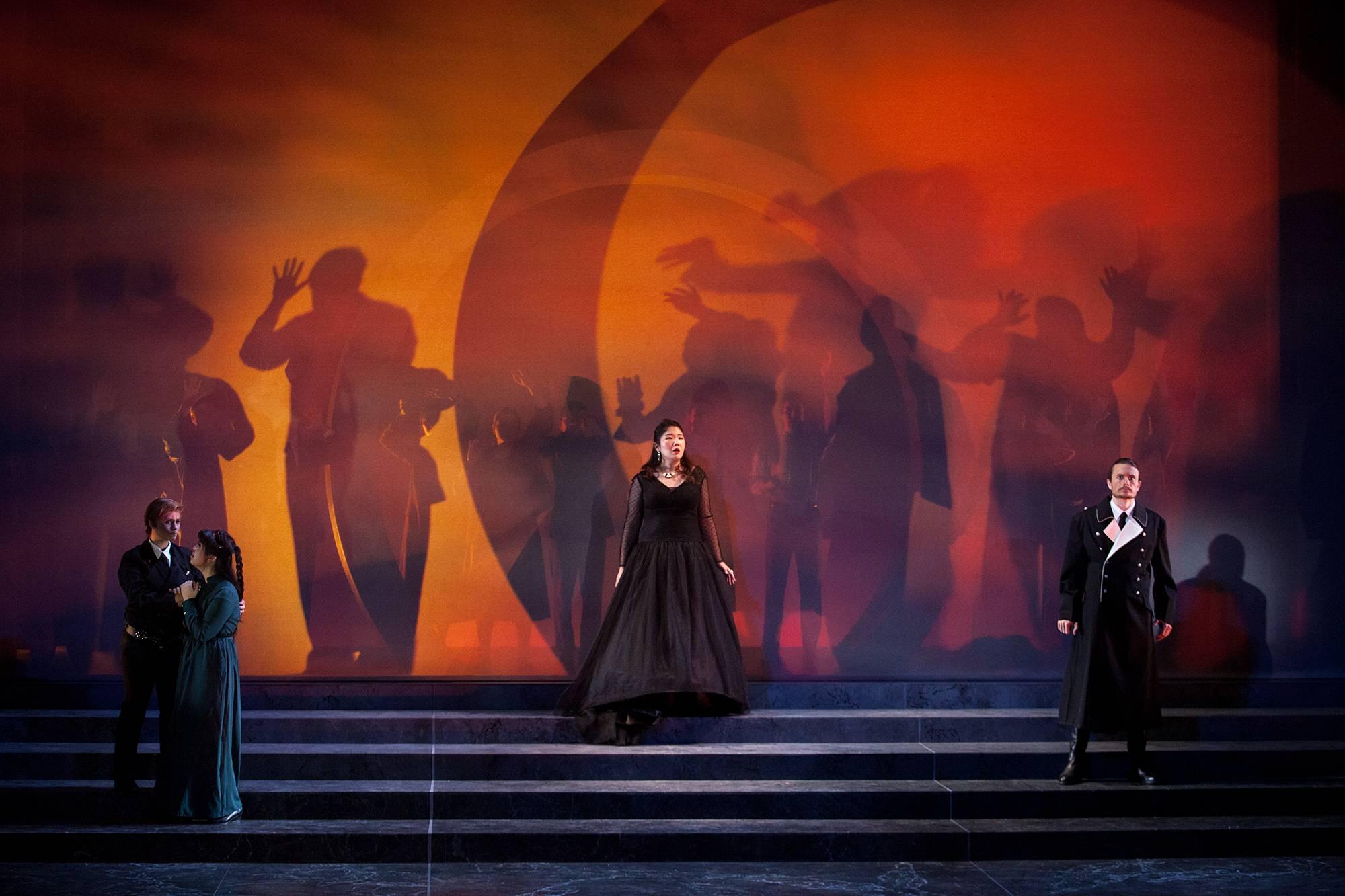 Three performers stand, spread across a wide, smoke-filled staircase, behind them is a spooky orange and red background with shadowy human figures projected against it. 