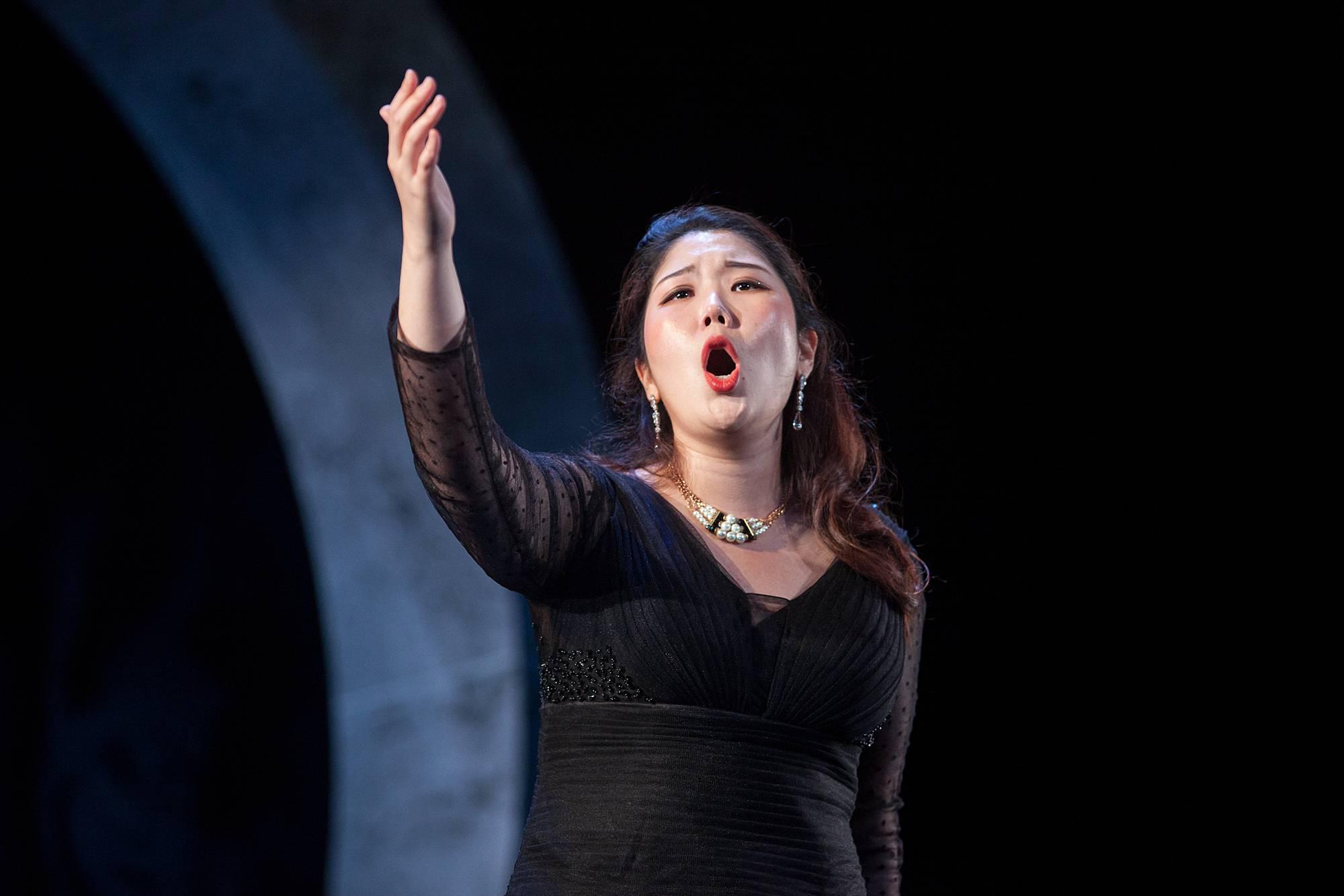 a woman sings in front of a stark, dark set, with one hand lifted toward the heavens. 