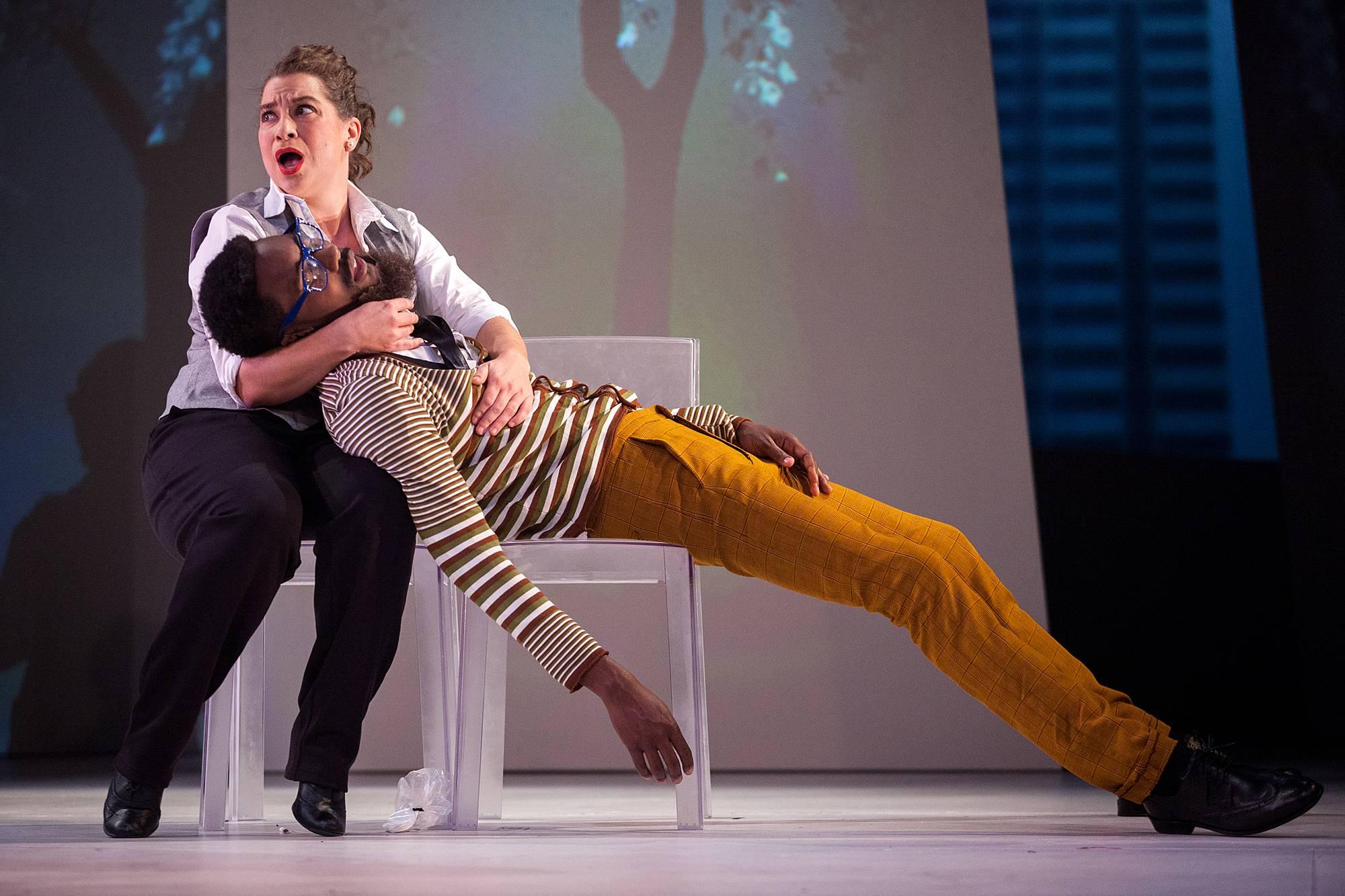 a woman sits in a chair on stage, holding a man in her arms who appears to be unconscious