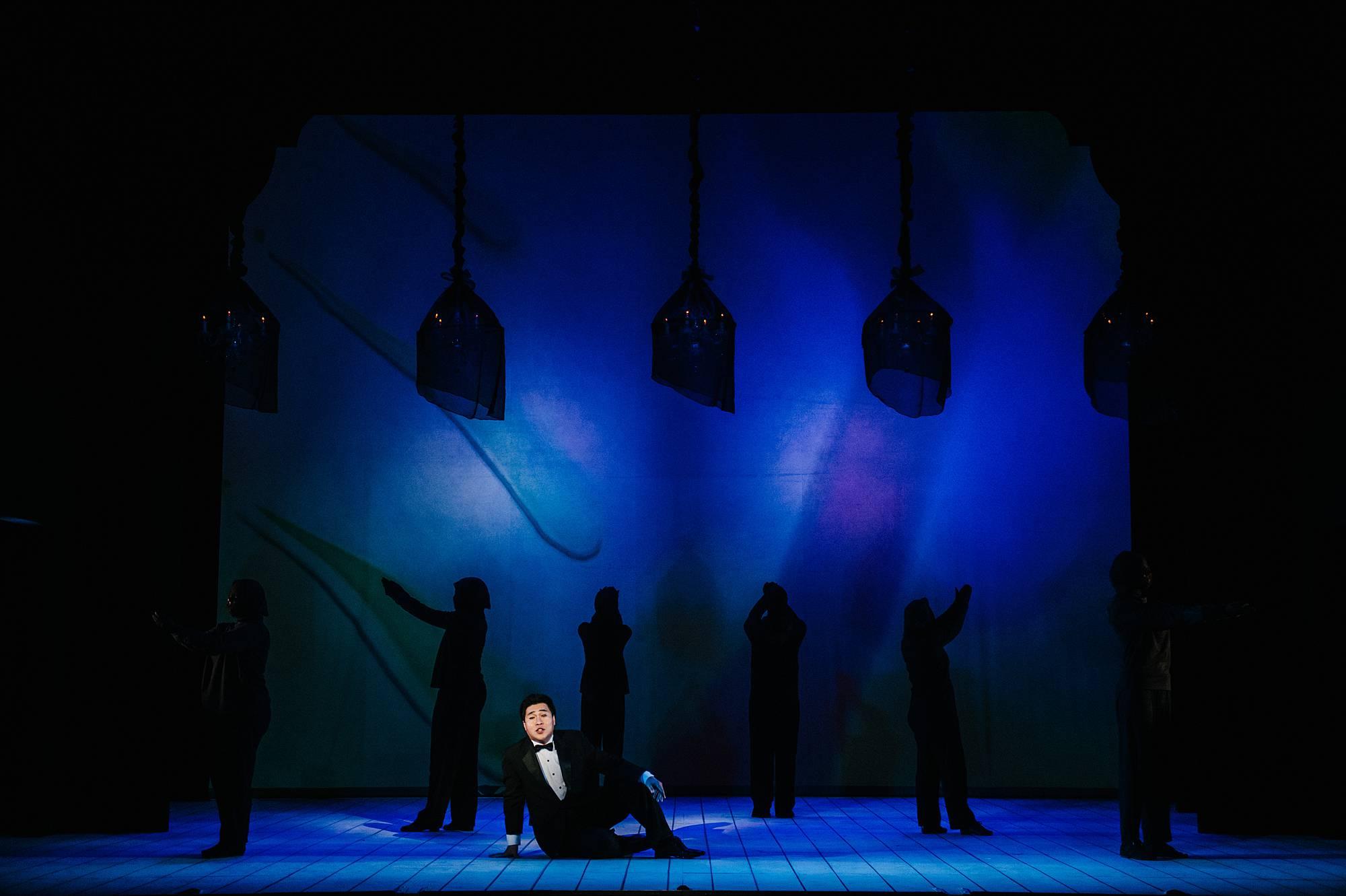 A singer in a tuxedo lounges causally on the stage floor, behind him shadowy figures in all black dance on a stage lit in deep blue light.