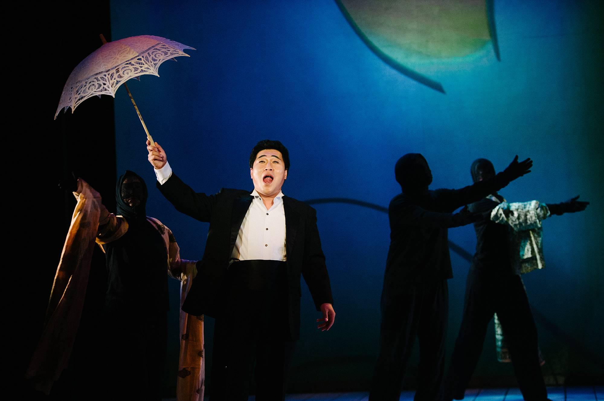 a Man in a tuxedo holds an open umbrella over his head as he sings, behind in shadowy figures dance.