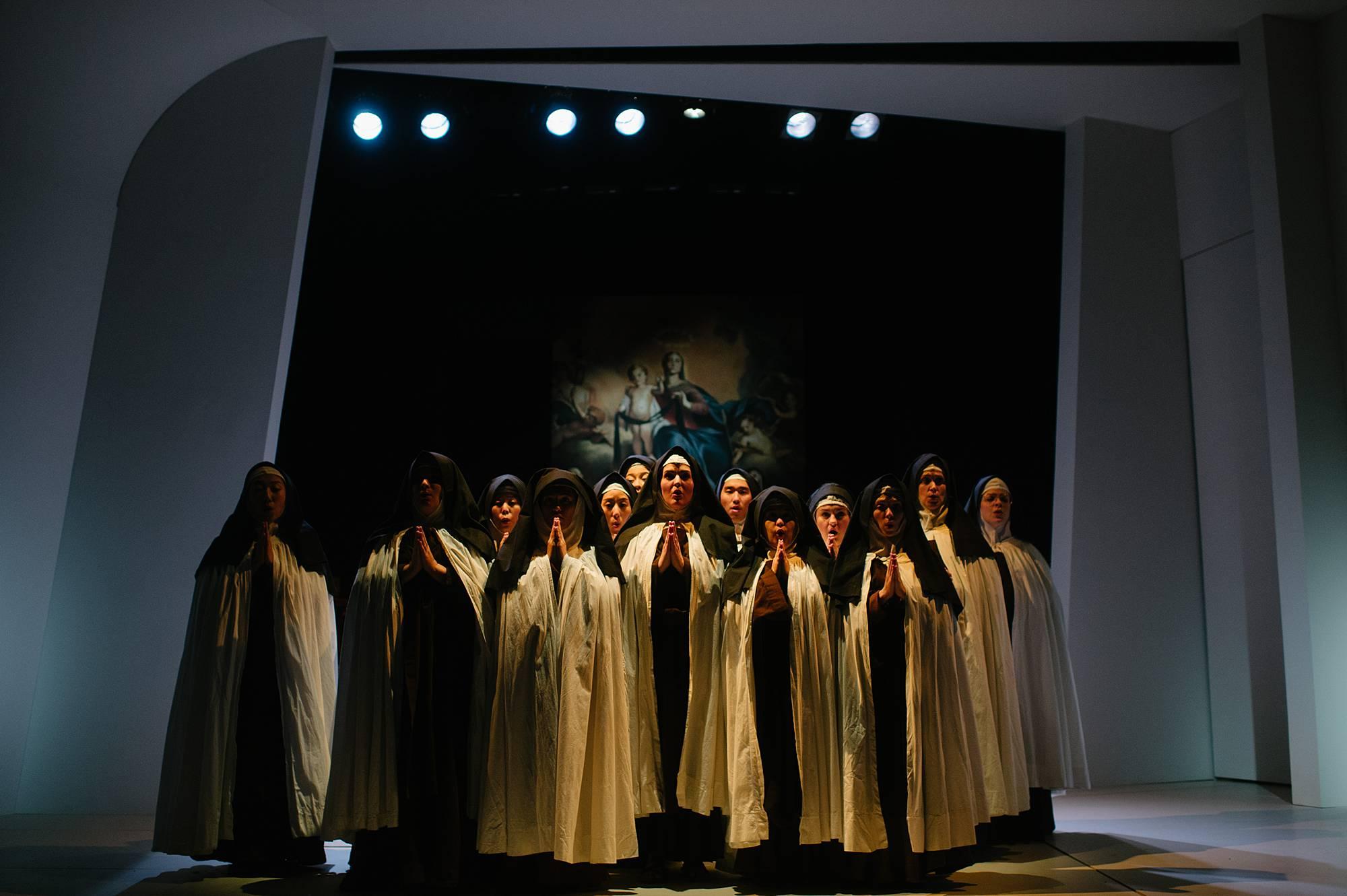 a group of nuns huddle together in dark light, hands clasped in prayer, looking up to the heavens.