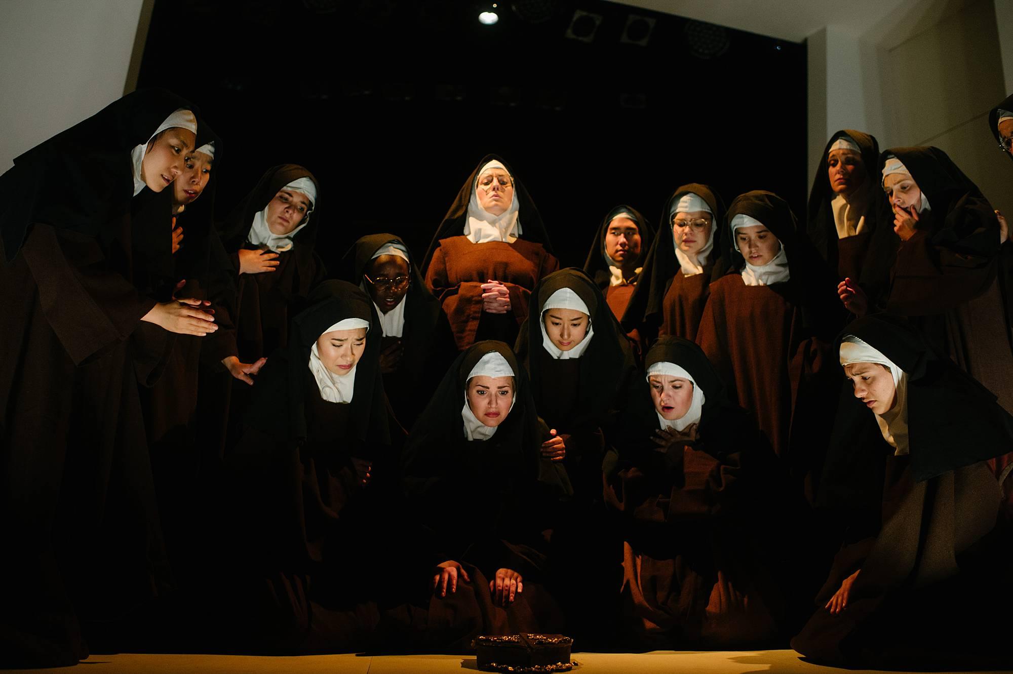 Three tiers of nuns kneel and stand over each other looking at a small music box in the foreground.