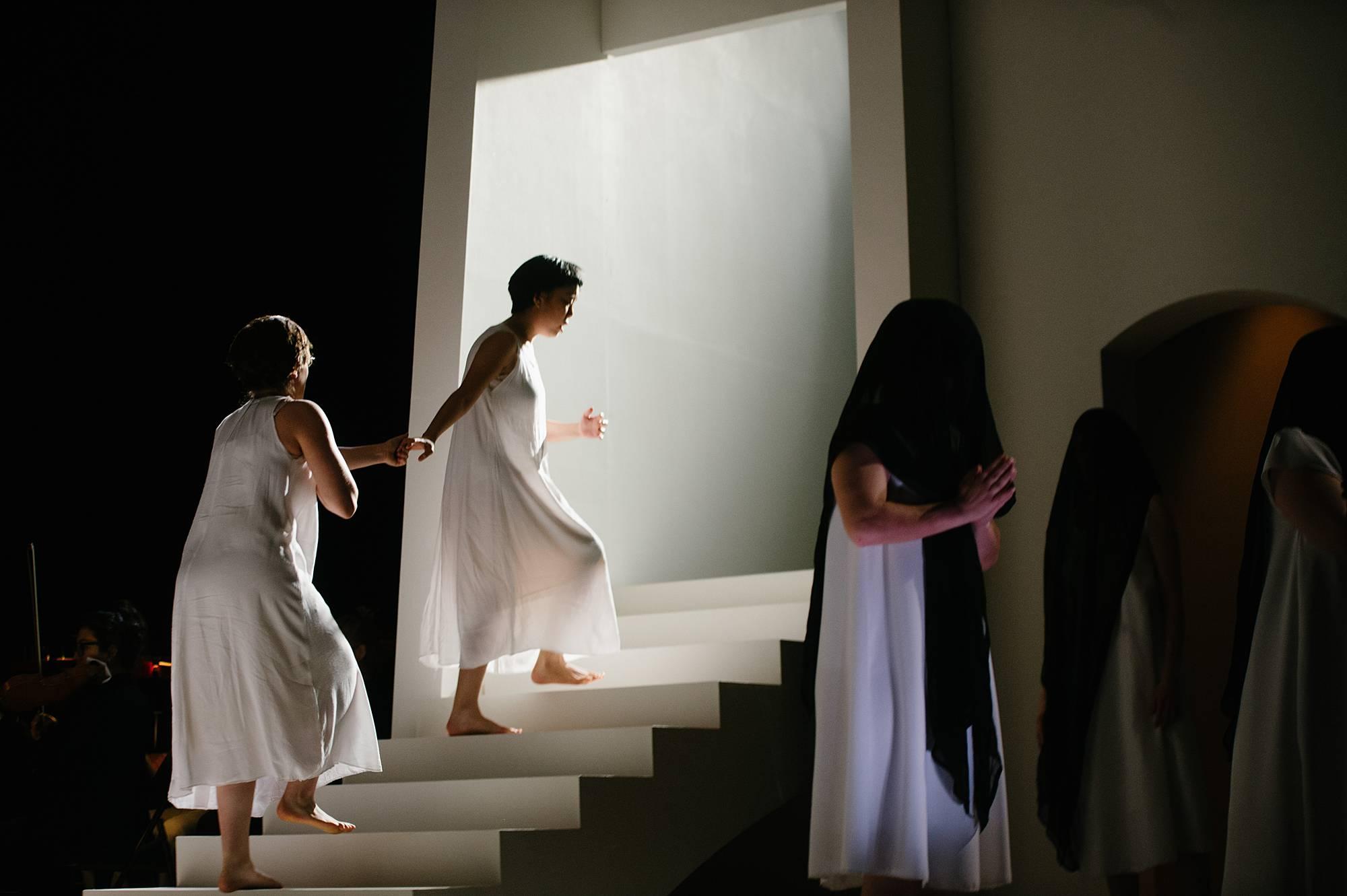 Two nuns wear only their undergarments. One leads the other up a staircase toward a bight white light, as if ascending into heaven.