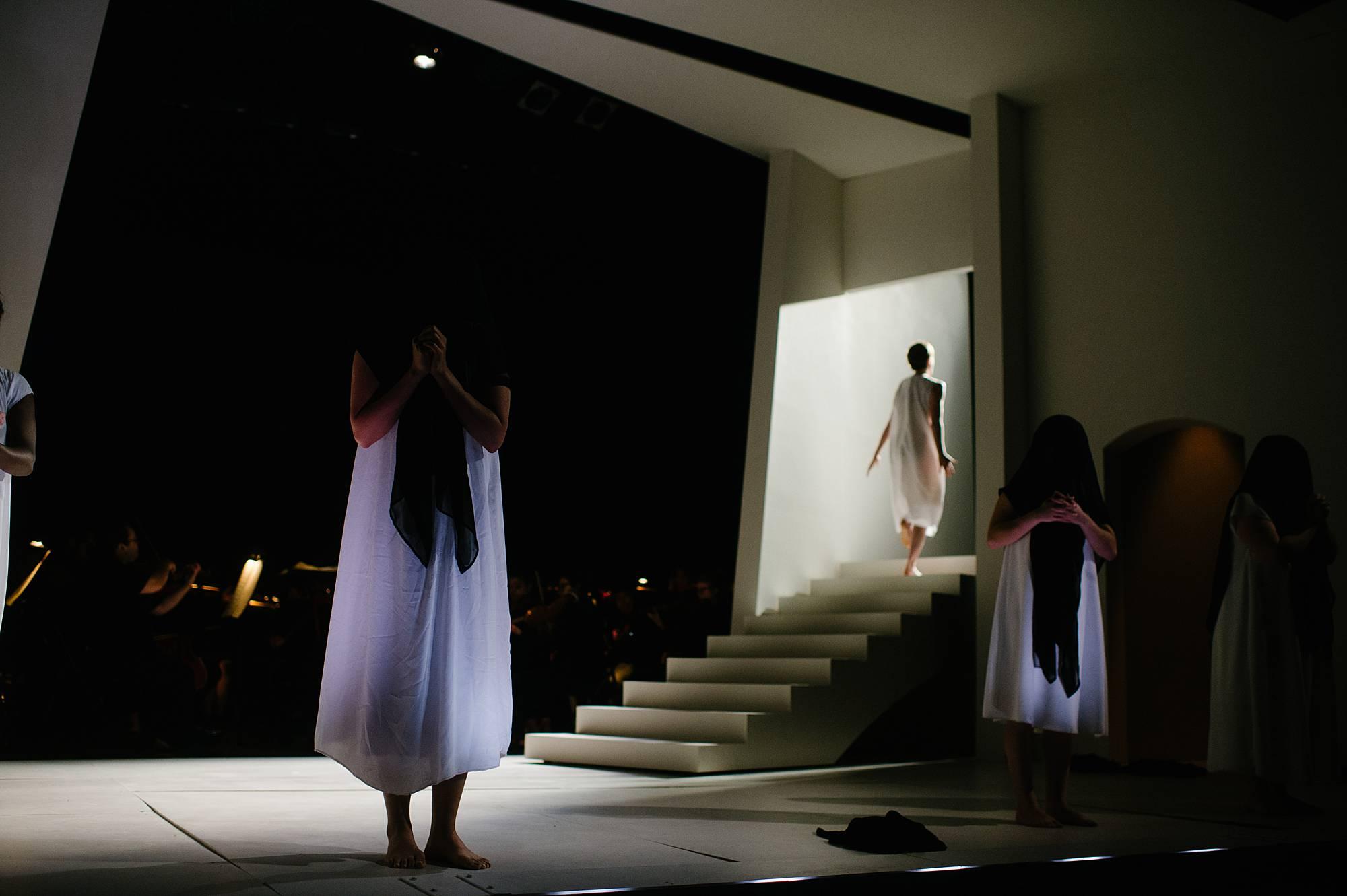 In the foreground a few figures in white gowns stand very still, their heads covered in black shawls.  In the background, out of focus, the final nun ascends a staircase into heaven. 