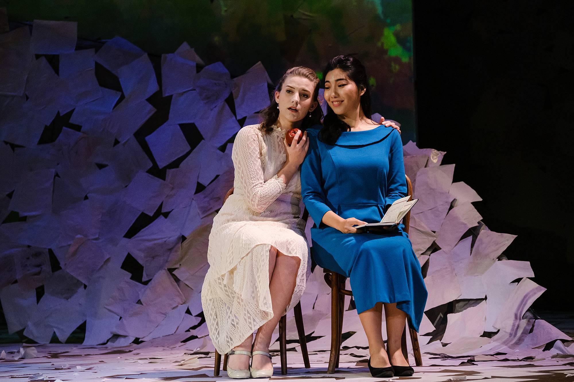 two women sit on small chairs center stage, holding each other close, as if telling one another secrets.