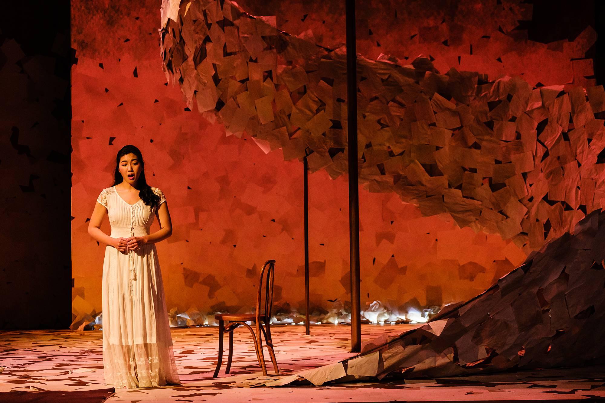 A woman in a white dress stands on a stage covered in paper, lit up in muted red tones.