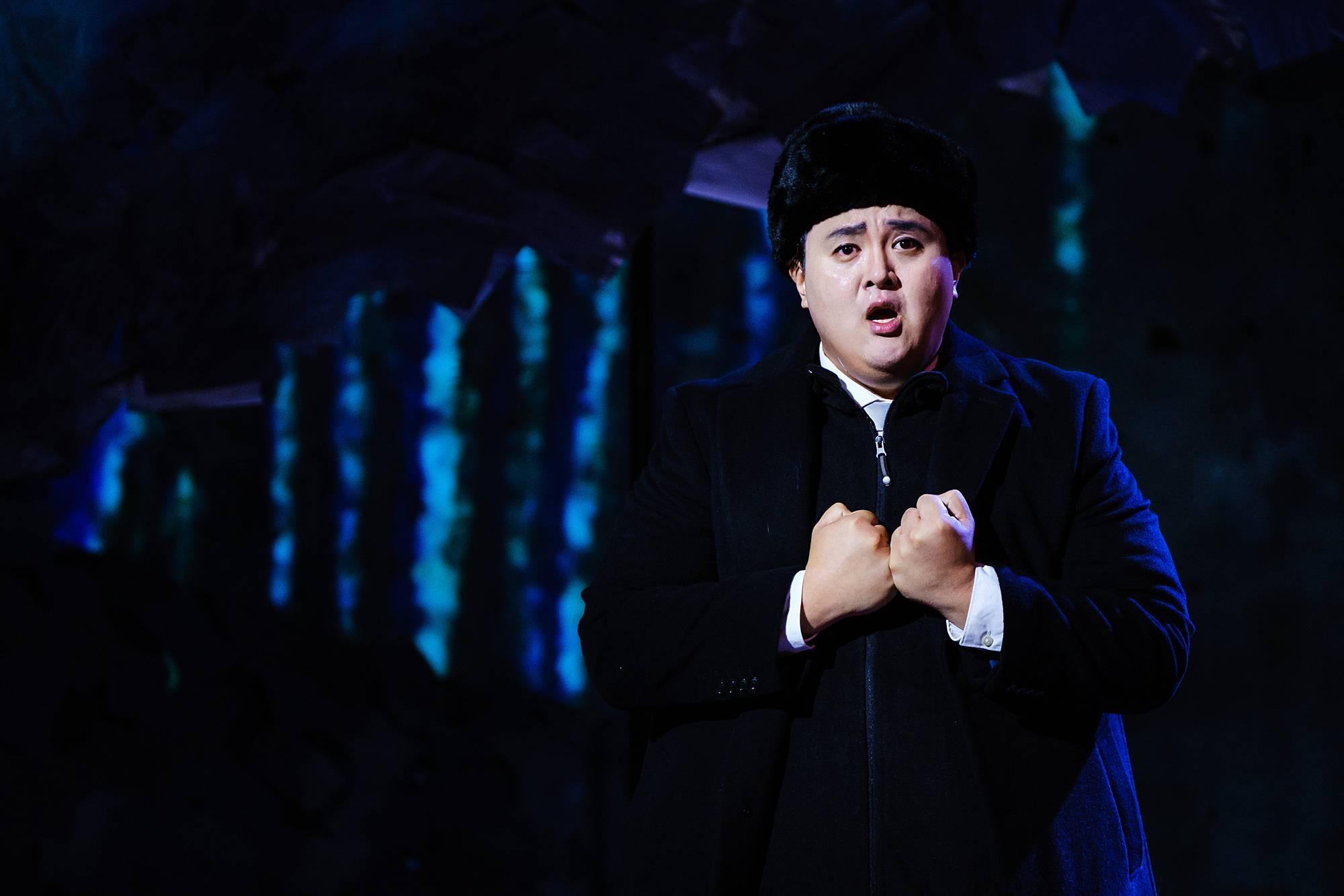 a man in an overcoat and bowler's hat stands stage right, with clenched fits, singing earnestly against a dark, night-time set.