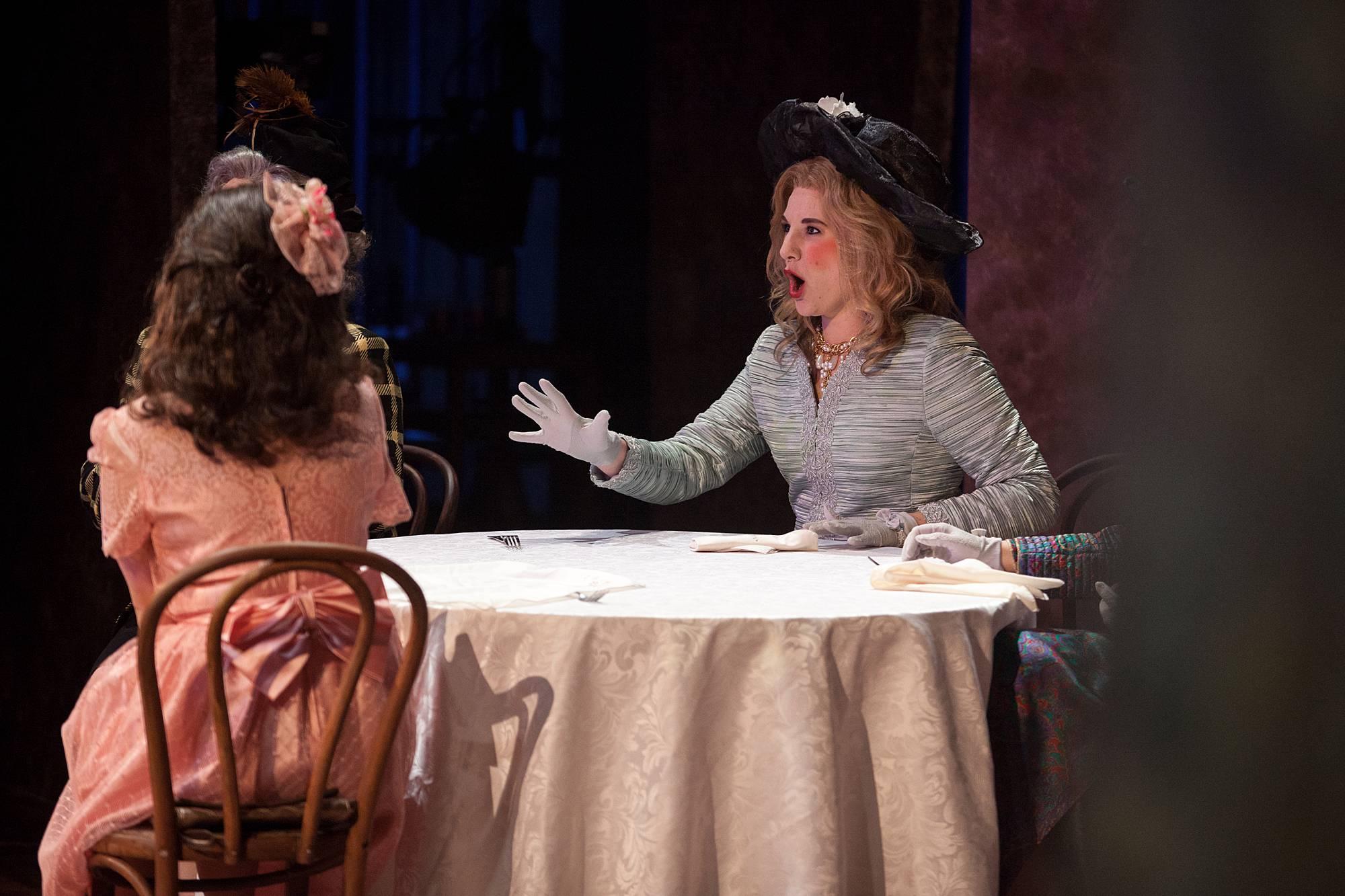 two women sit at a bar table having an argument. One woman's back is turned to the audience.