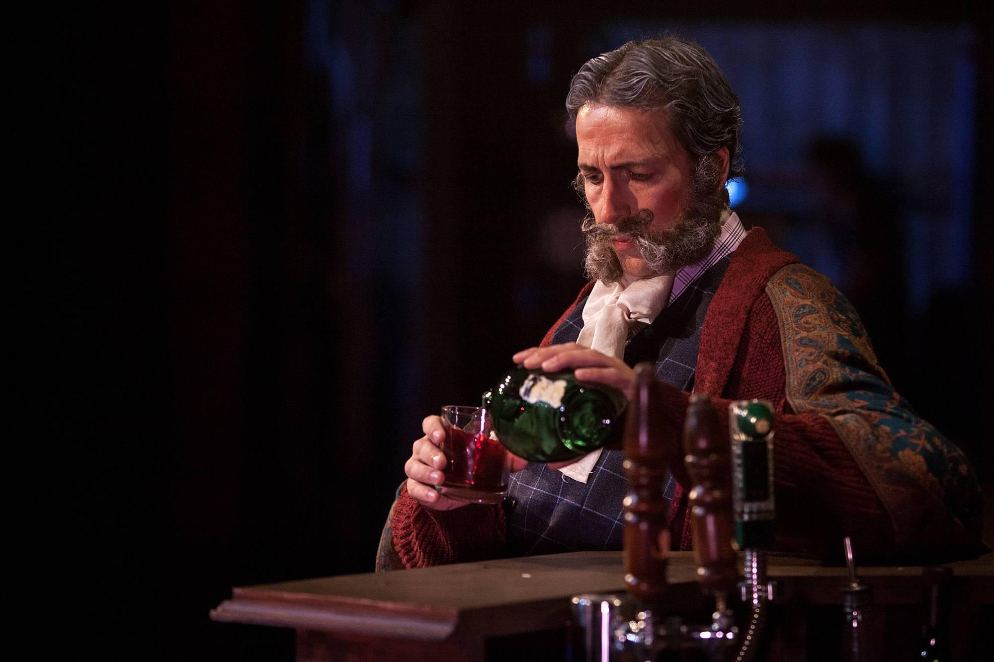 An elderly man with a neatly trimmed beard sits a table and pours himself a drink.