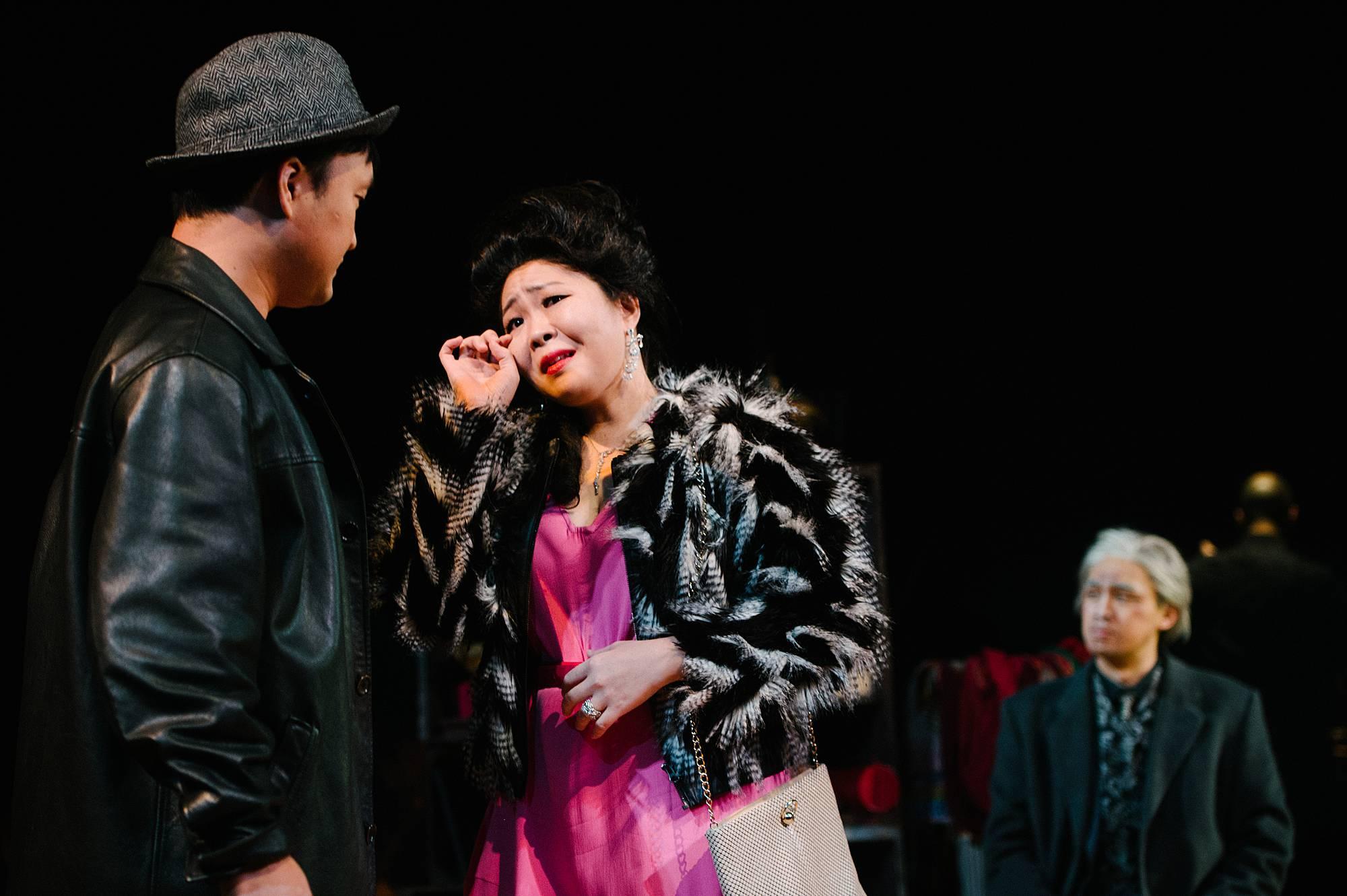 a woman in a bright pink dress and overcoat speaks tearfully with a man wearing a Fedora hat. 