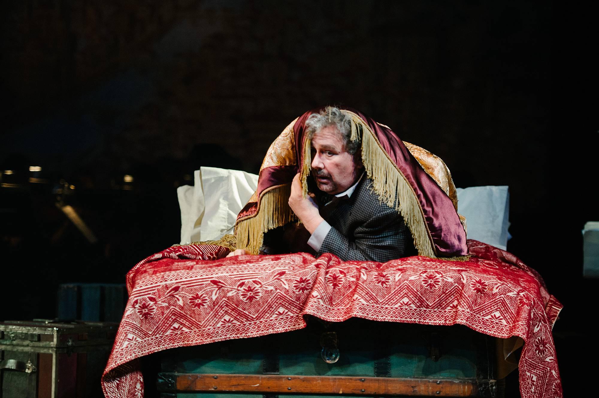 a man in a suit and tie lies on a bed, covered loosely by a comforter, his head is at the foot of the bed, and he peers out from the comforter as though he is hiding from someone. 