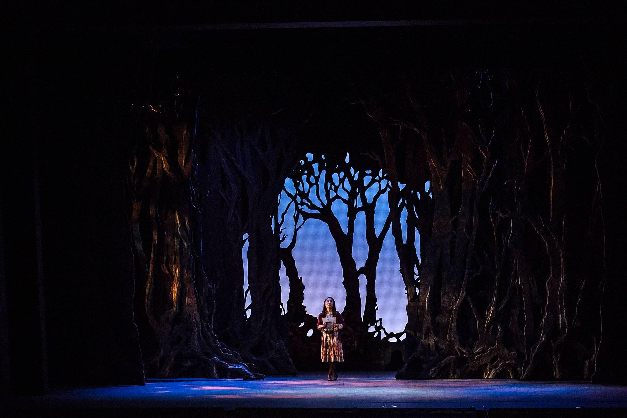 Gretel stands amid an epic forest set, holding a map and looking uncertain and frightened.