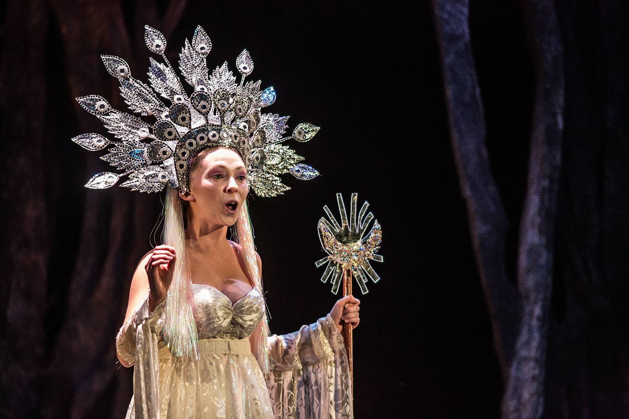 A woman stands dressed in white, with an elaborate headdress and scepter