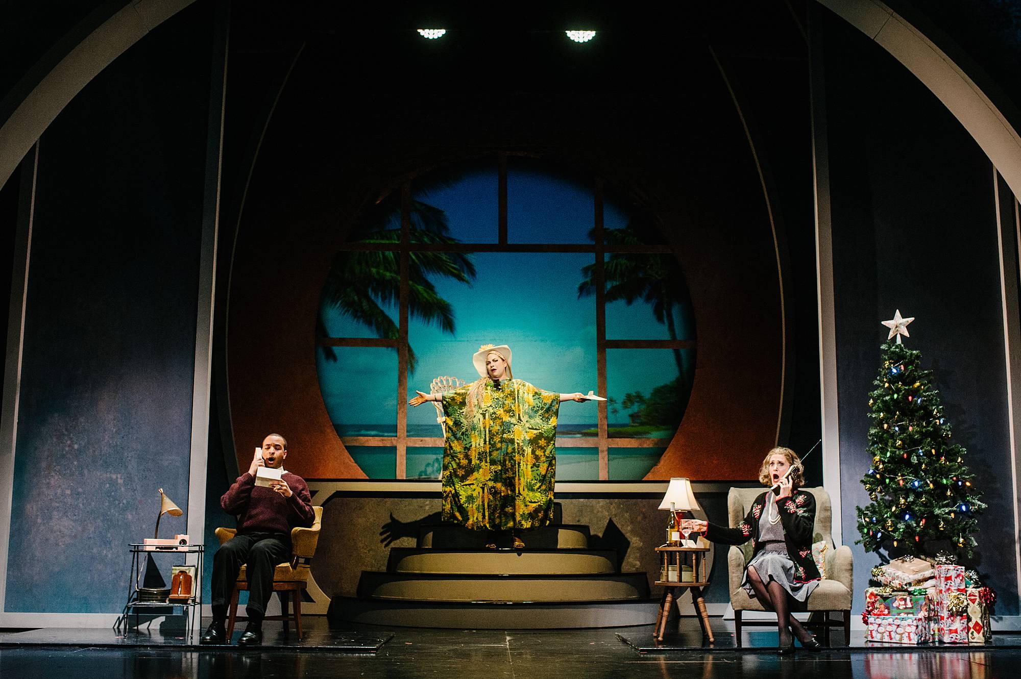 One opposites of the stage a woman and man sit, talking on phones. A Christmas tree is on the right side of the stage.  Center stage behind them a woman in a moo moo stands with arms outstretched in front of a projection of a tropical beach