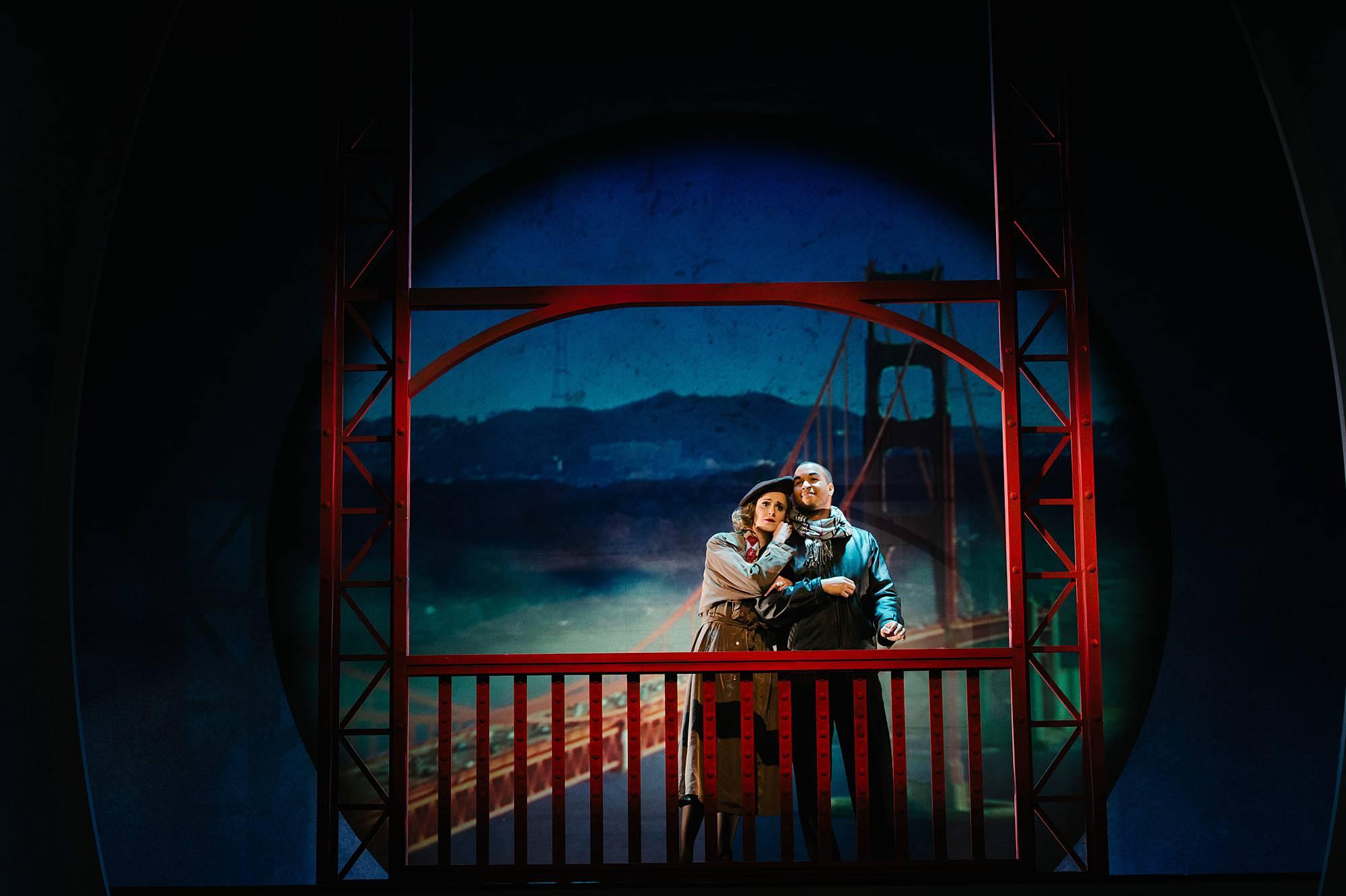 A woman cuddles up to a man, resting her head on his shoulder. They are standing in winter coats and scarves on the Golden Gate Bridge.