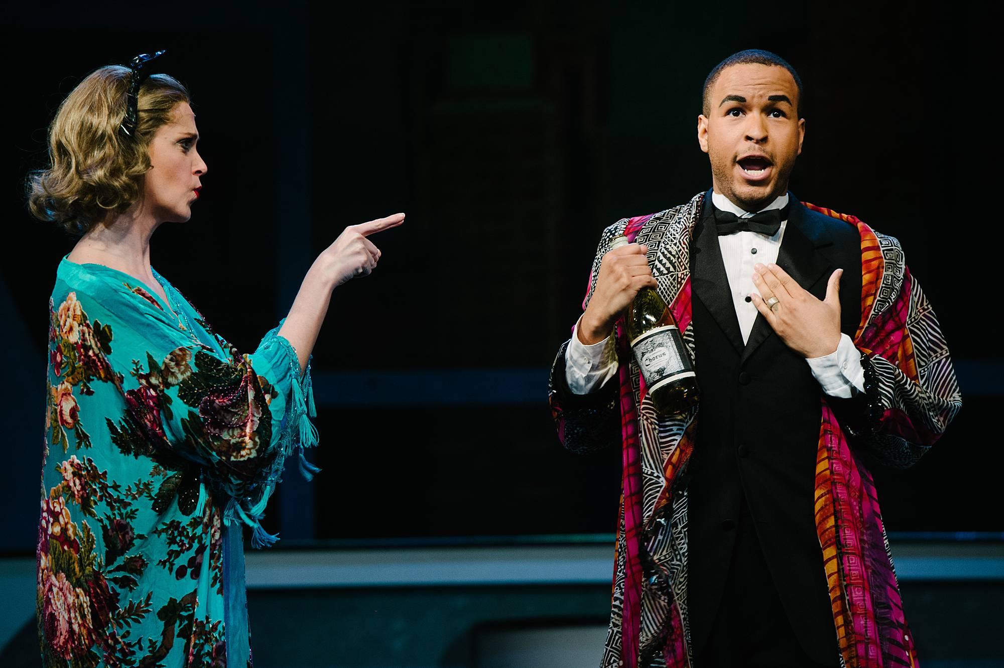 A woman in a silk robe points angrily at a man also in a silk robe, clutching a bottle of champagne. He looks surprised at the woman't accusation.