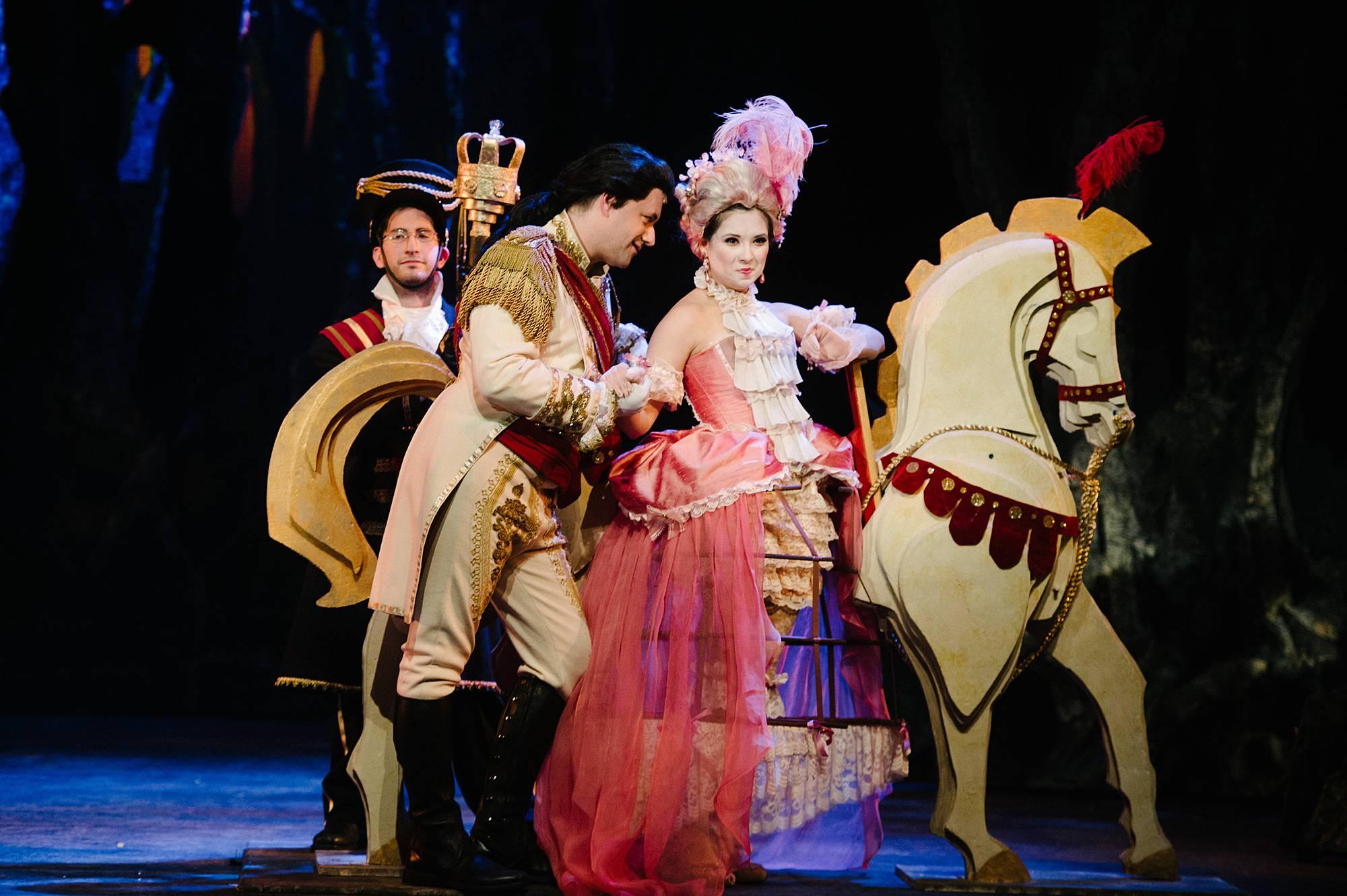 A woman in formal Elizabethan attire ride atop a white steed, while she is fawned over by a suitor in coat and tails.