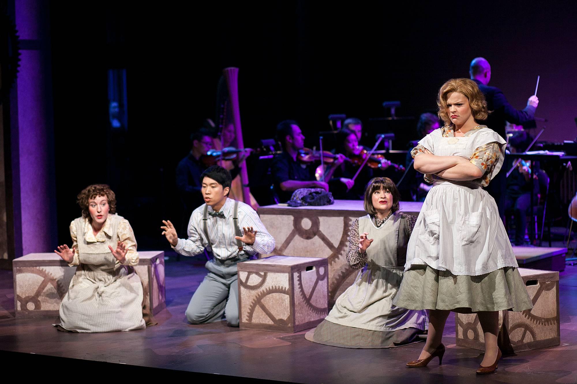 2 girls and a boy sit on their knees around stage, while another girl stands, arms crossed and an angry scowl on her face.