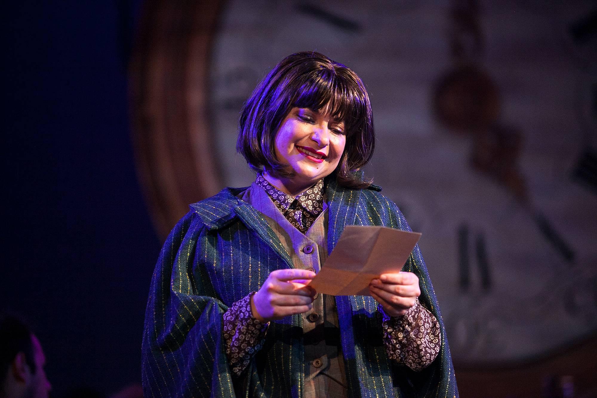 An older woman stands center stage, and reads a letter with a sweet smile on her face.