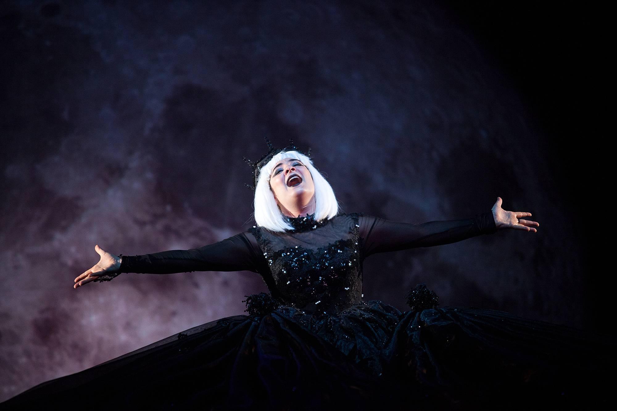 A woman in with stark white hair in a black dress lifts her hands and face skyward on stage. 