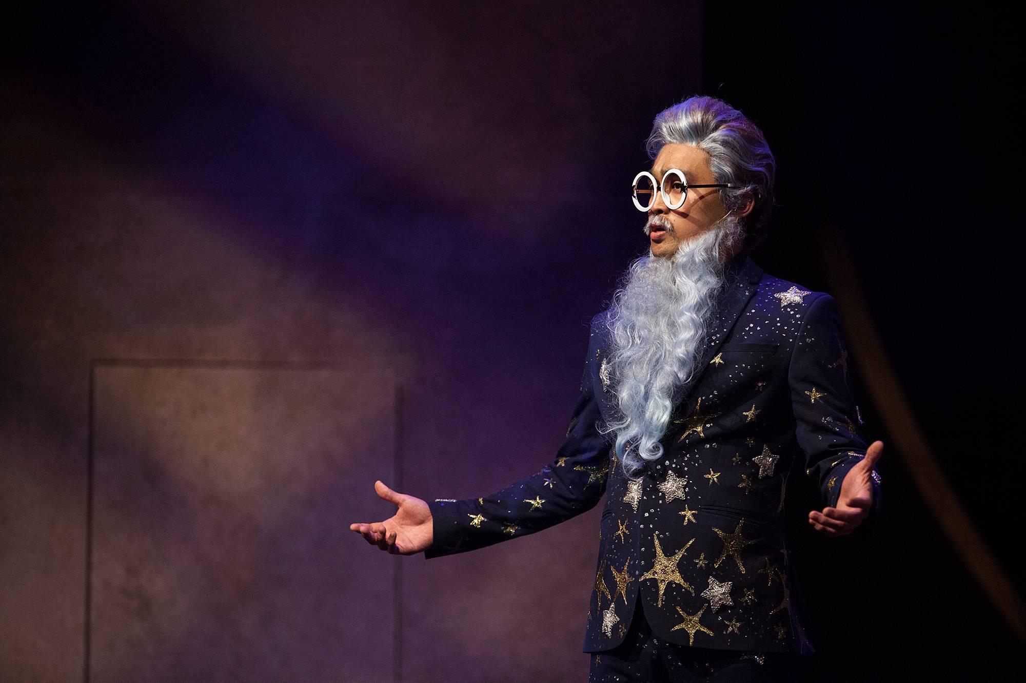 A man with a long white beard wearing oversized round spectacles and a suit covered in falling stars sings with arms outstretched.