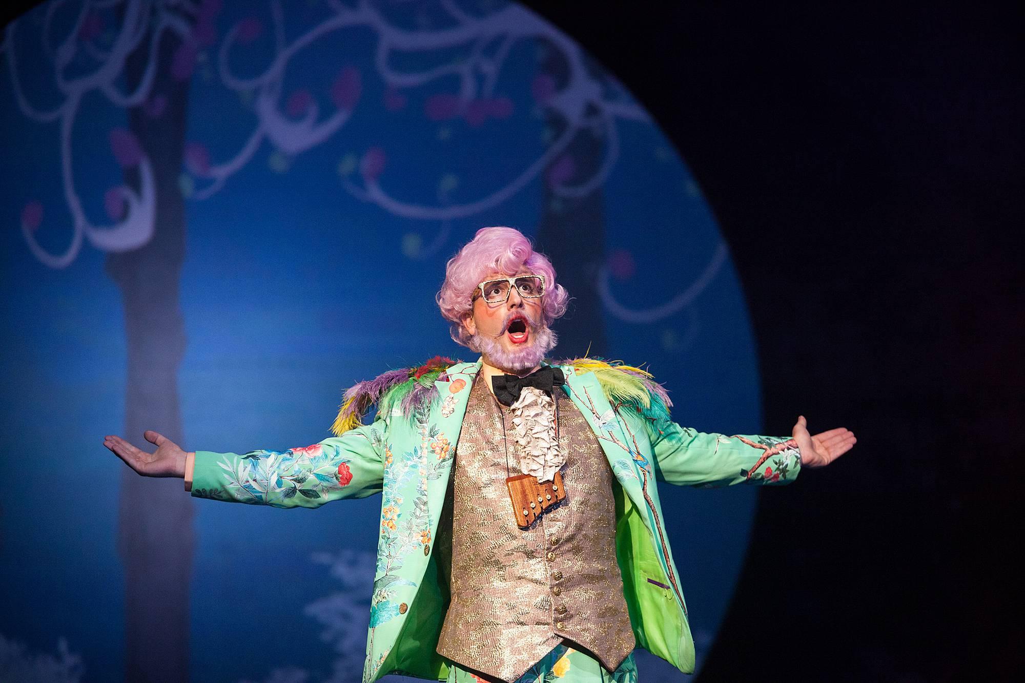 a man with a pink bouffant and beard wearing a bright green suit and waistcoat sings with arms outstretched.