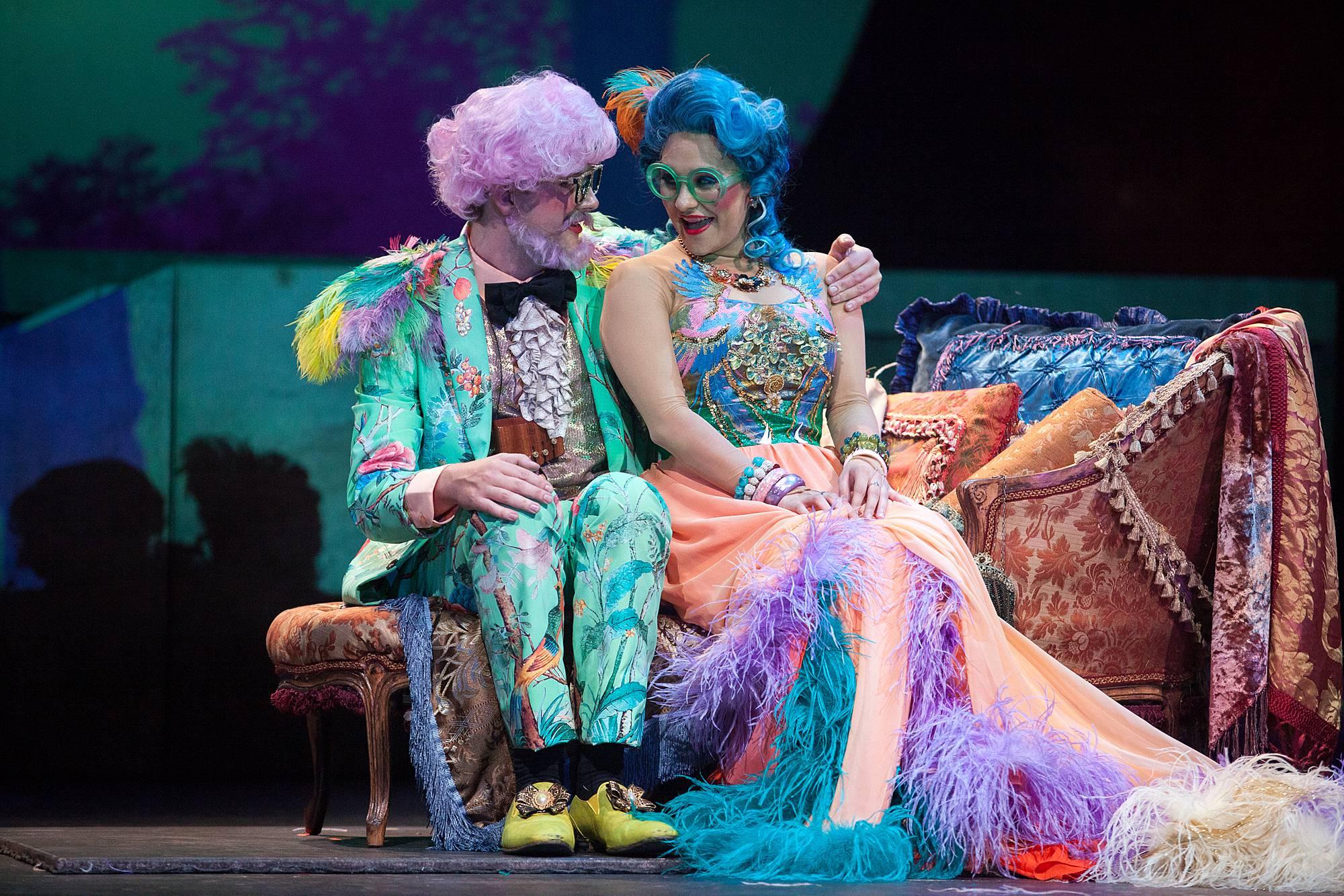 a man with a pink bouffant and beard dressed in a bright green suit sits on a couch with his arm wrapped around a woman with blue hair and a very colorful dress.