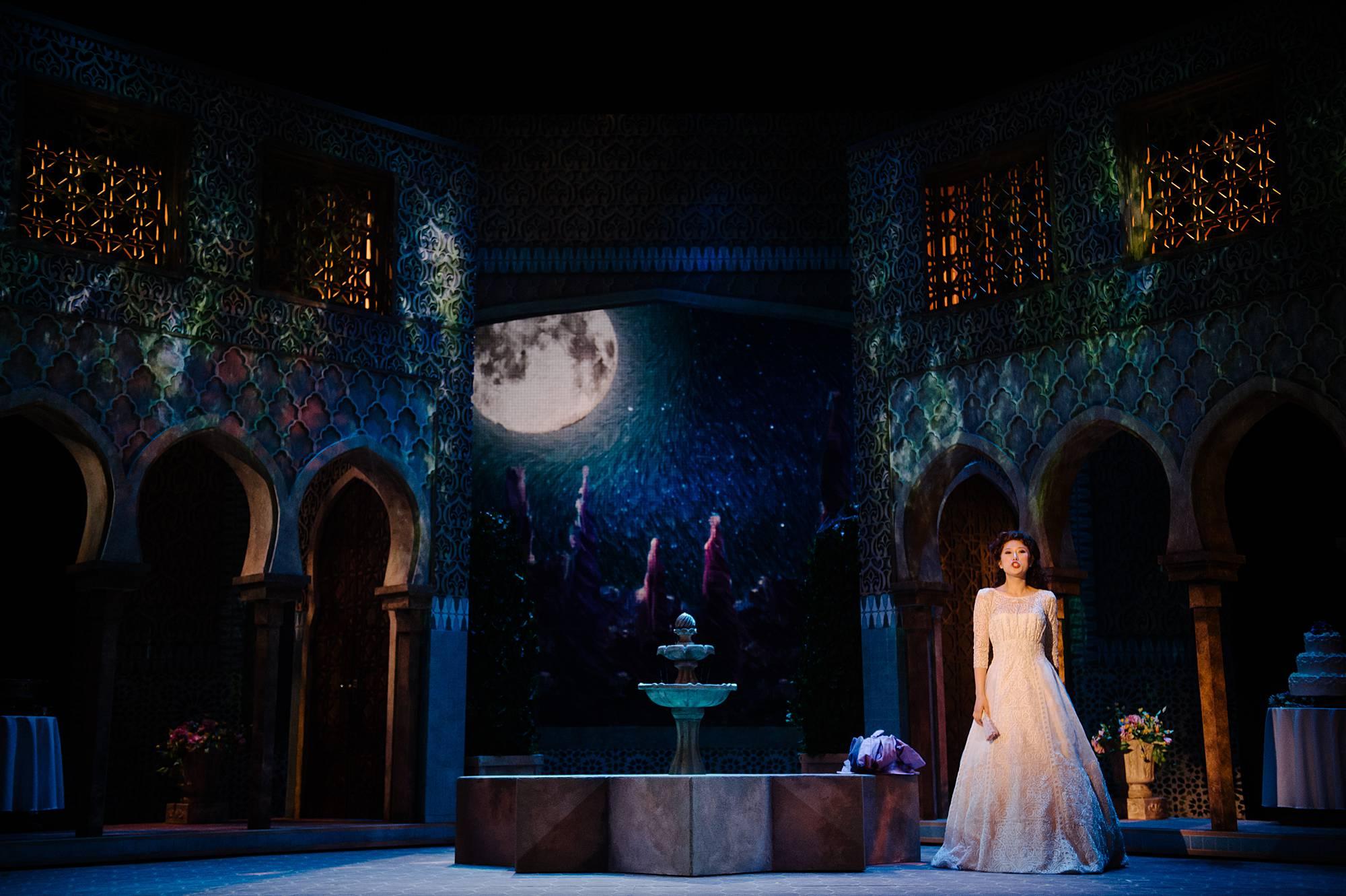 A woman in a wedding gown stands alone by a fountain at night, a soft moon is projected behind her. 